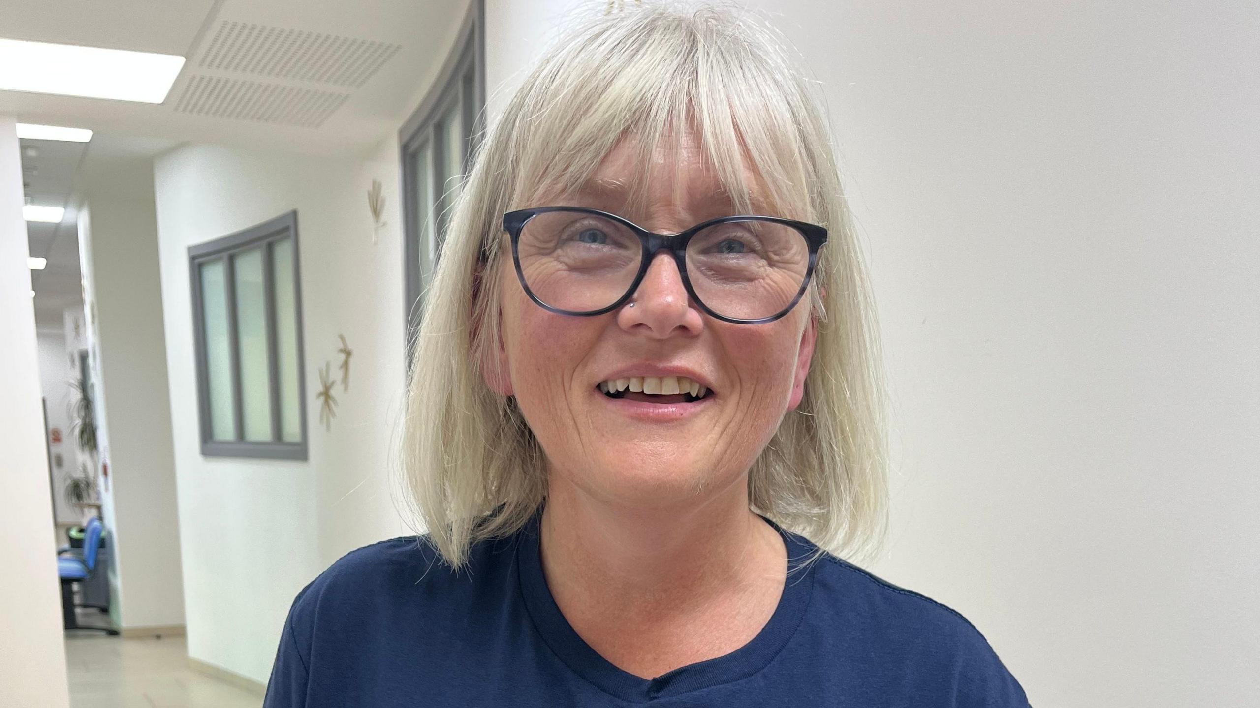 A woman with black glasses and a grey bob haircut is smiling to the camera. She is wearing a blue T-shirt with "Mouth Cancer Foundation" on it.