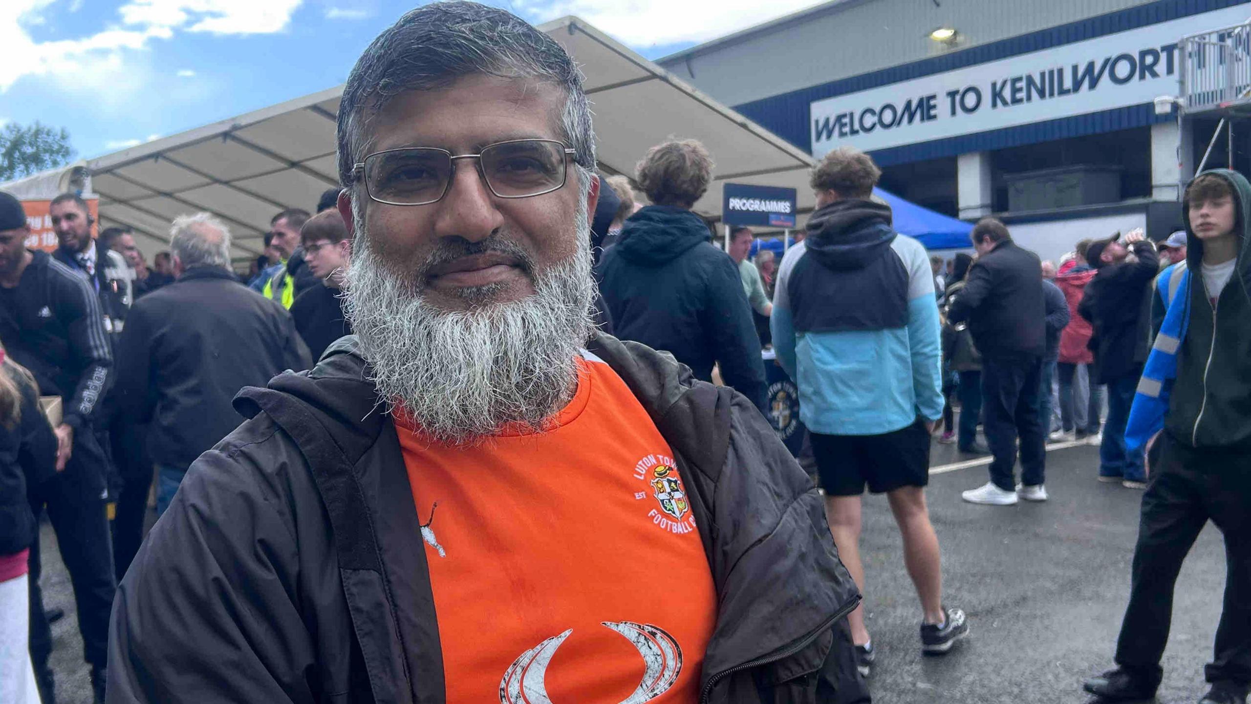 Abdul Ghafoor wears an orange Luton Town T-shirt and a black jacket. He has black short hair and a grey beard and is also wearing glasses. He is standing in front of football fans and the stadium, with a sign that says 'Welcome to Kenilworth...'.