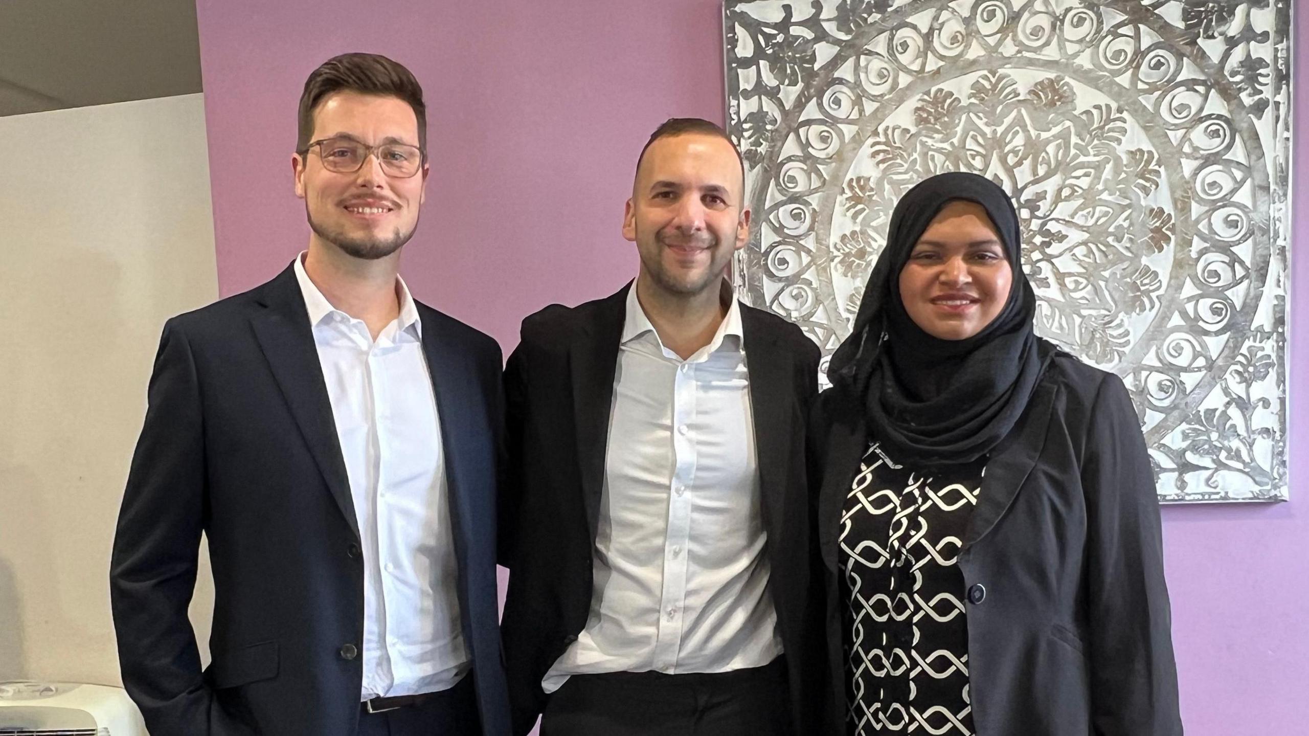 Three people stand close together smiling at the camera. Two men, Tom Butcher and Zack Polanski, are in black suits with open-neck shirts, while Repi Begum is on the right, wearing a black blazer with a patterned top.
