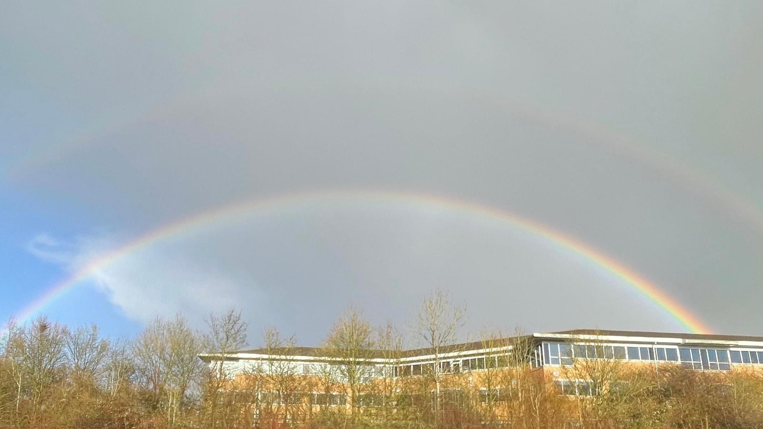 A double rainbow in a mostly grey sky, some blue peaking through on the left. They sit above an office building with some trees in front.