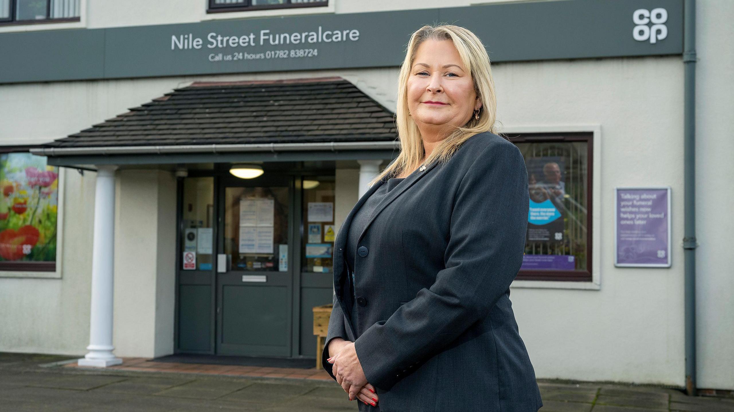 A woman wearing a black suit stands and looks into the camera. A building with the words Nile Street Funeralcare is behind her.