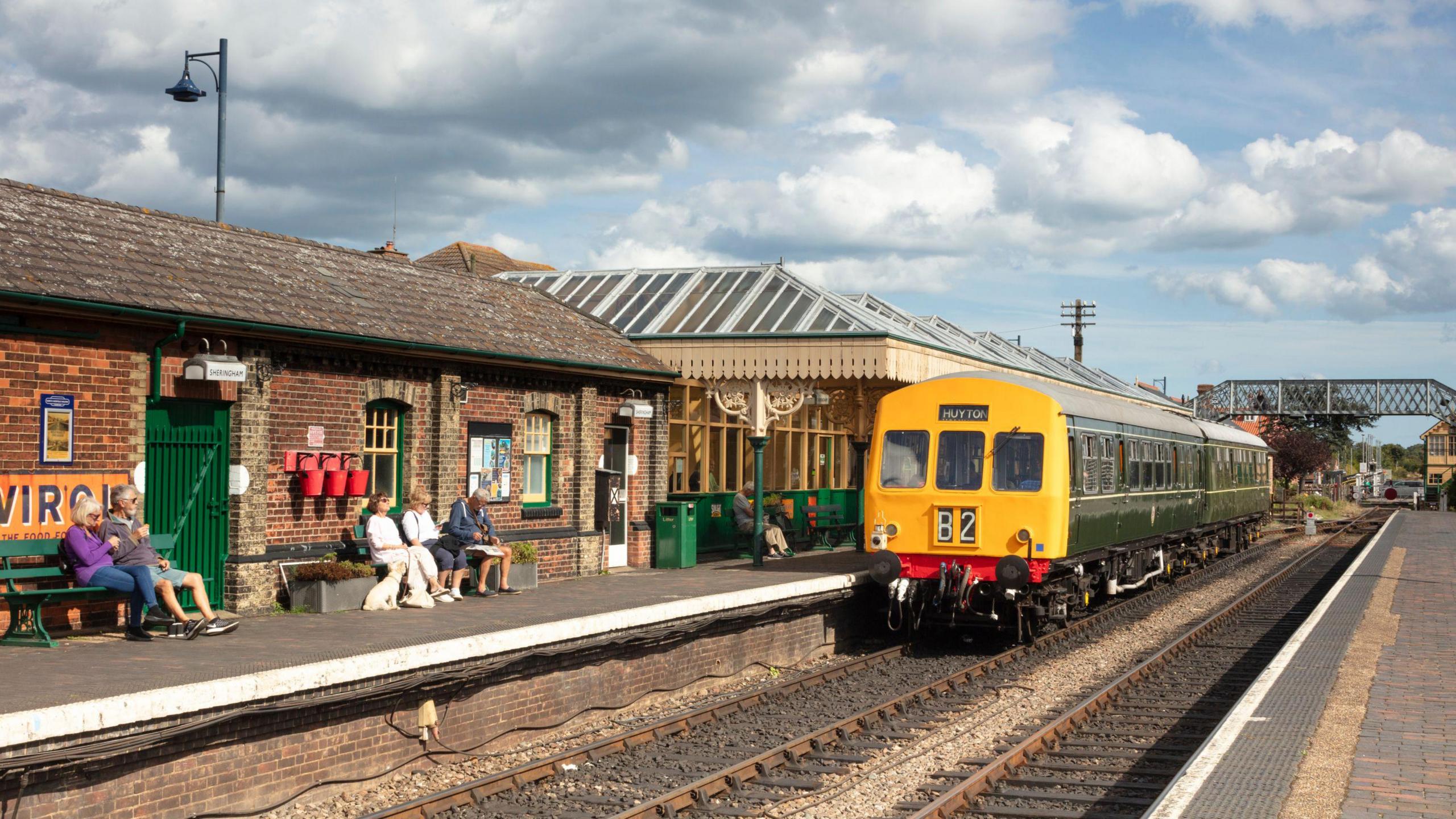 Six people sit on the green benches of a heritage rail station as a yellow and green two-coach train sits alongside the platform.