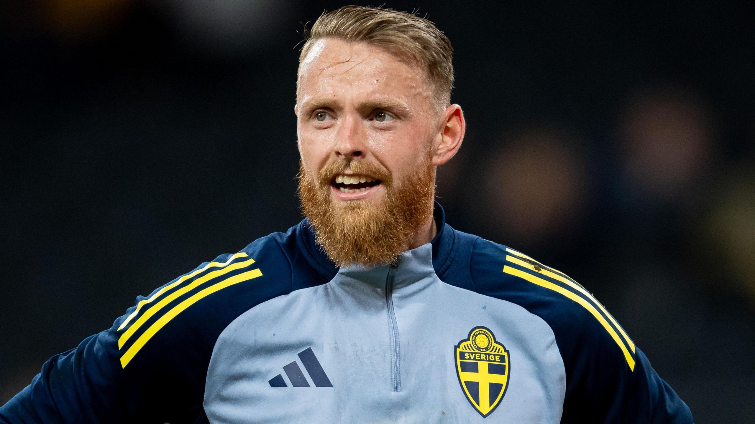Viktor Johansson wearing a light blue tracksuit top with navy blue shoulders and sleeves and yellow Adidas stripes, and a Sweden badge on the front. He can be seen smiling.
