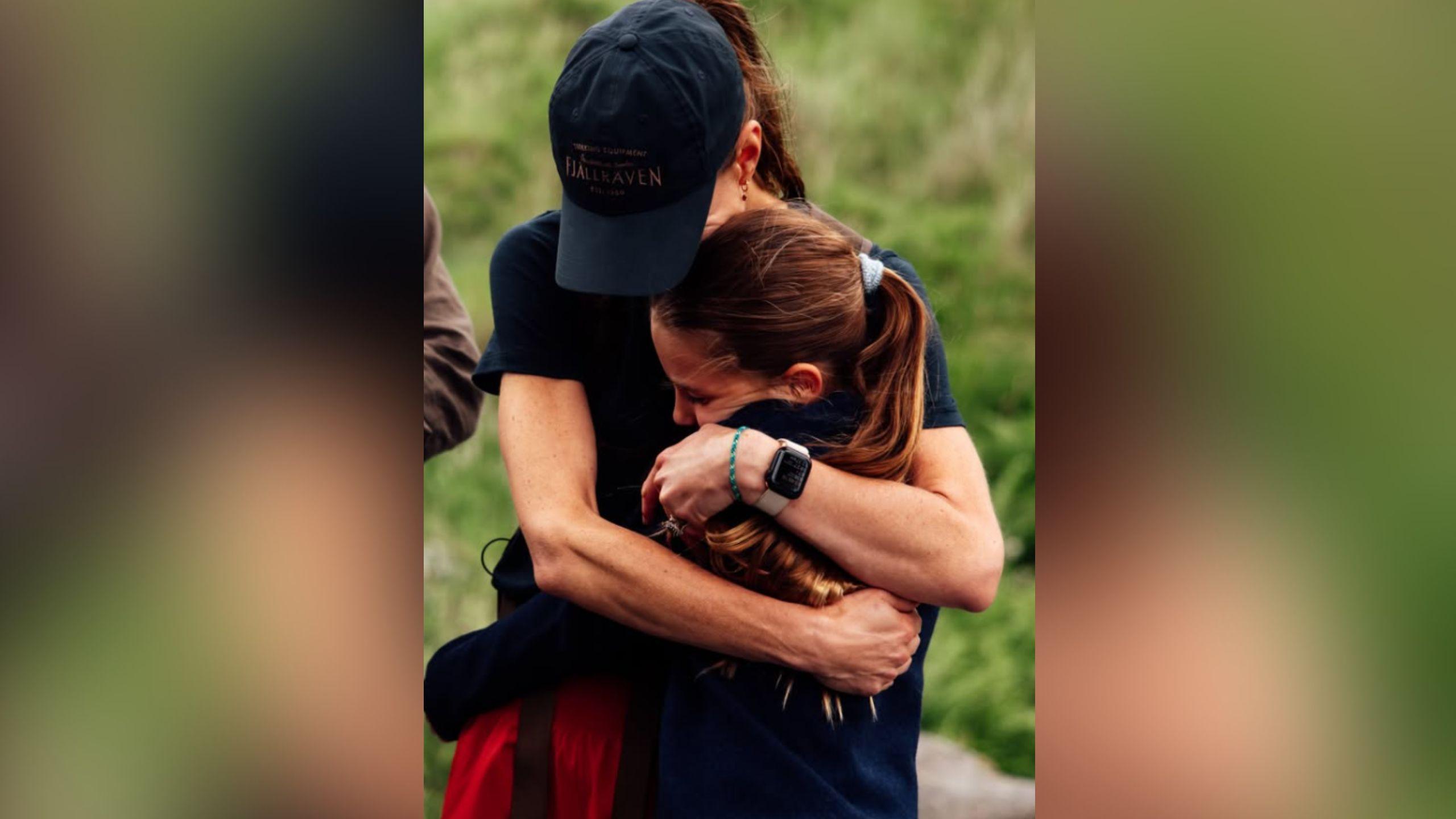Catherine hugs her daughter Charlotte. Catherine wears a black baseball cap, a black t-shirt and red shorts. Charlotte has long brown hair tied up in a ponytail. Behind them is a green hillside.