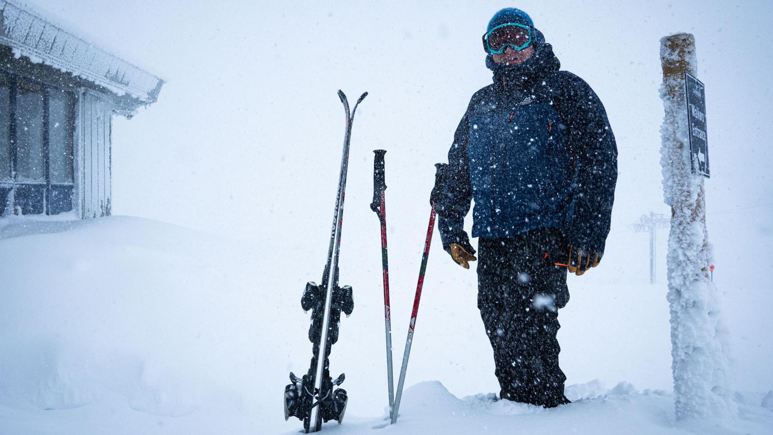 A skier stands in snow next to a pair of skis and ski poles which are stuck into the snow. The skier is wearing a fleece hat, goggles, a thick blue coat and black ski trousers.