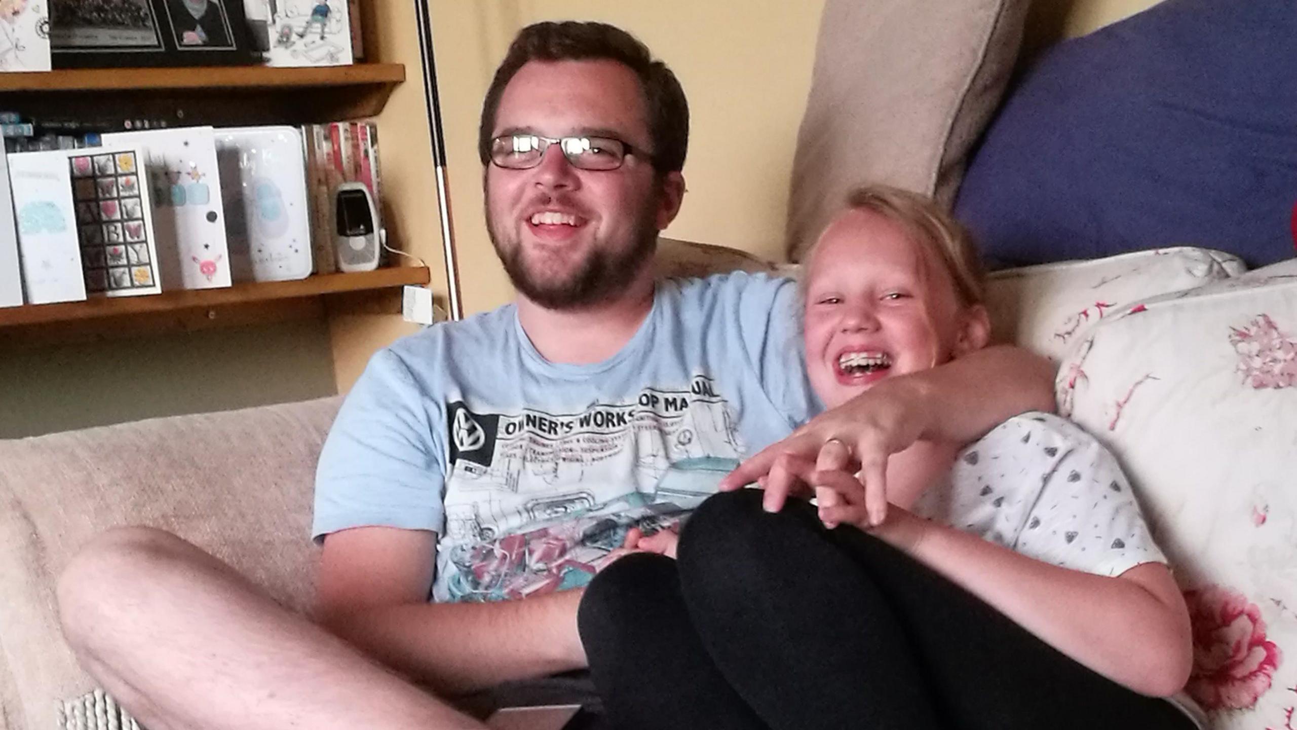 A man with short brown hair, stubble and rectangular glasses is wearing a blue top and smiling on a sofa with his arm around a teenager. The teenager has her mouth open laughing with braces and has blonde hair tied back, a white top and black leggings. Their hands are entwined. Next to them is a shelf with cards and books on it.