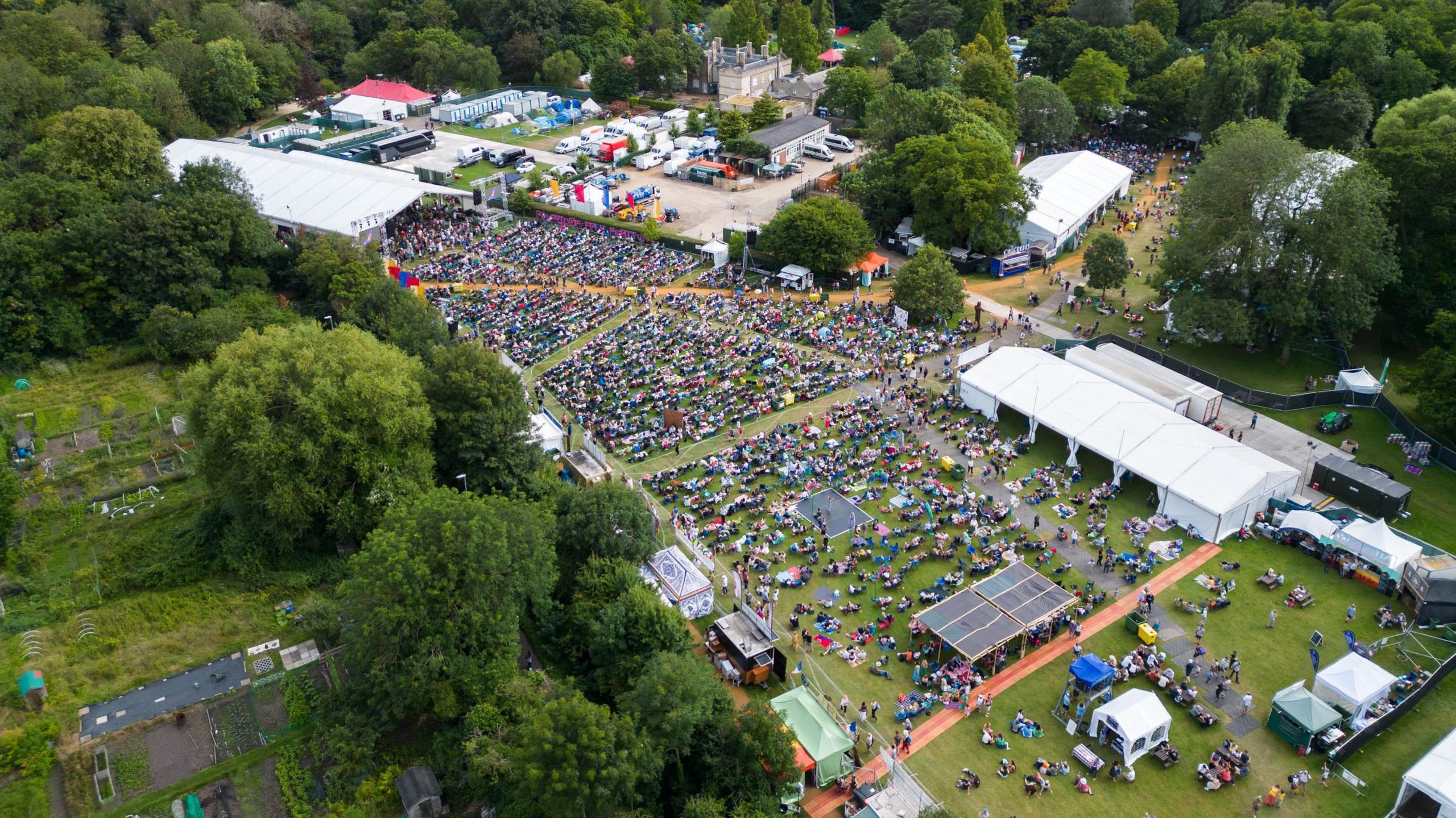 Aerial view of the folk festival in 2024. Three large white tents have been put up across fields and there are many people on the ground. People are watching performances at the stages and sat on the grass around them.