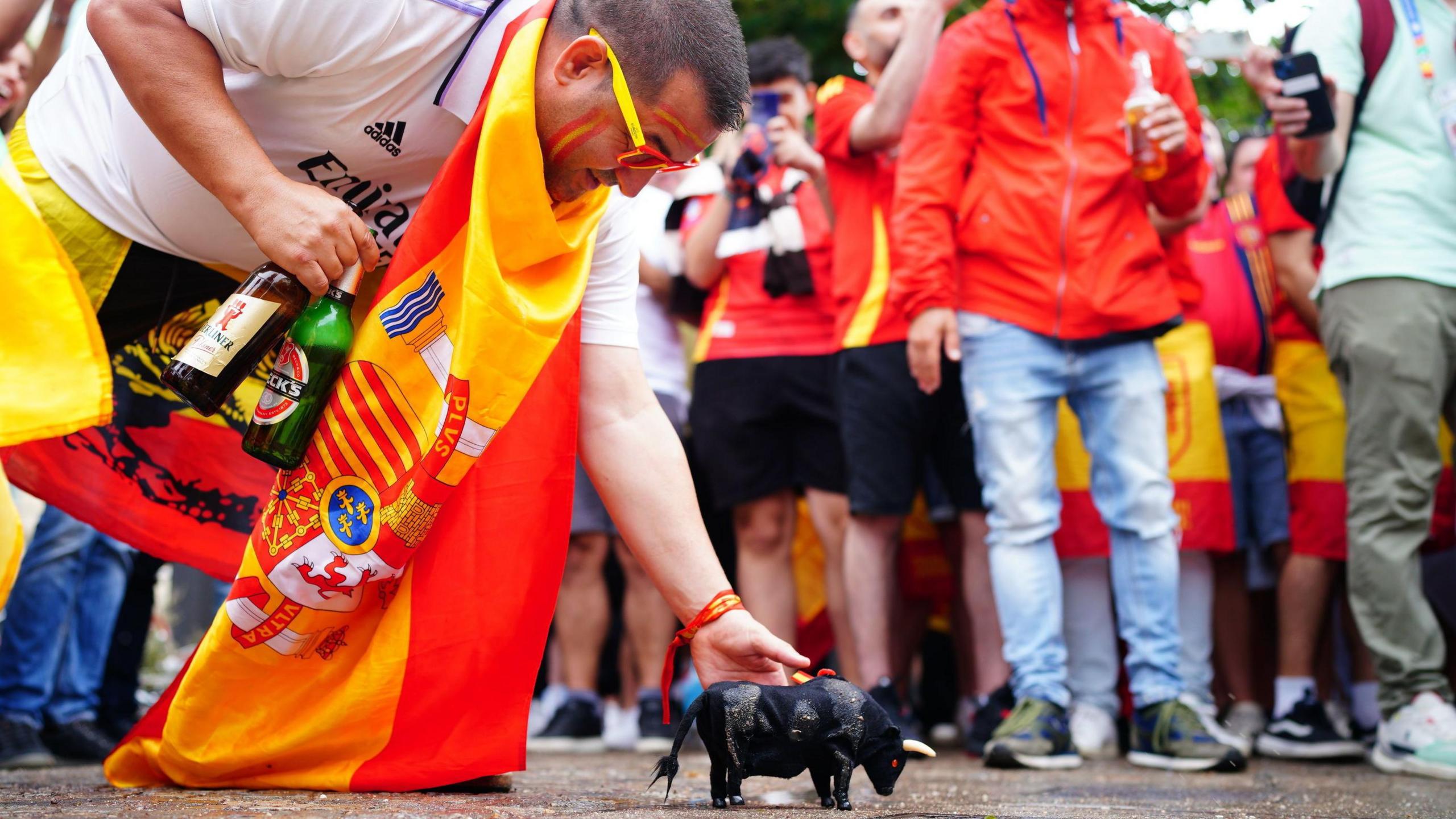 Man wearing Spanish scarf round his neck places a plastic bull on the ground in front of a crowd