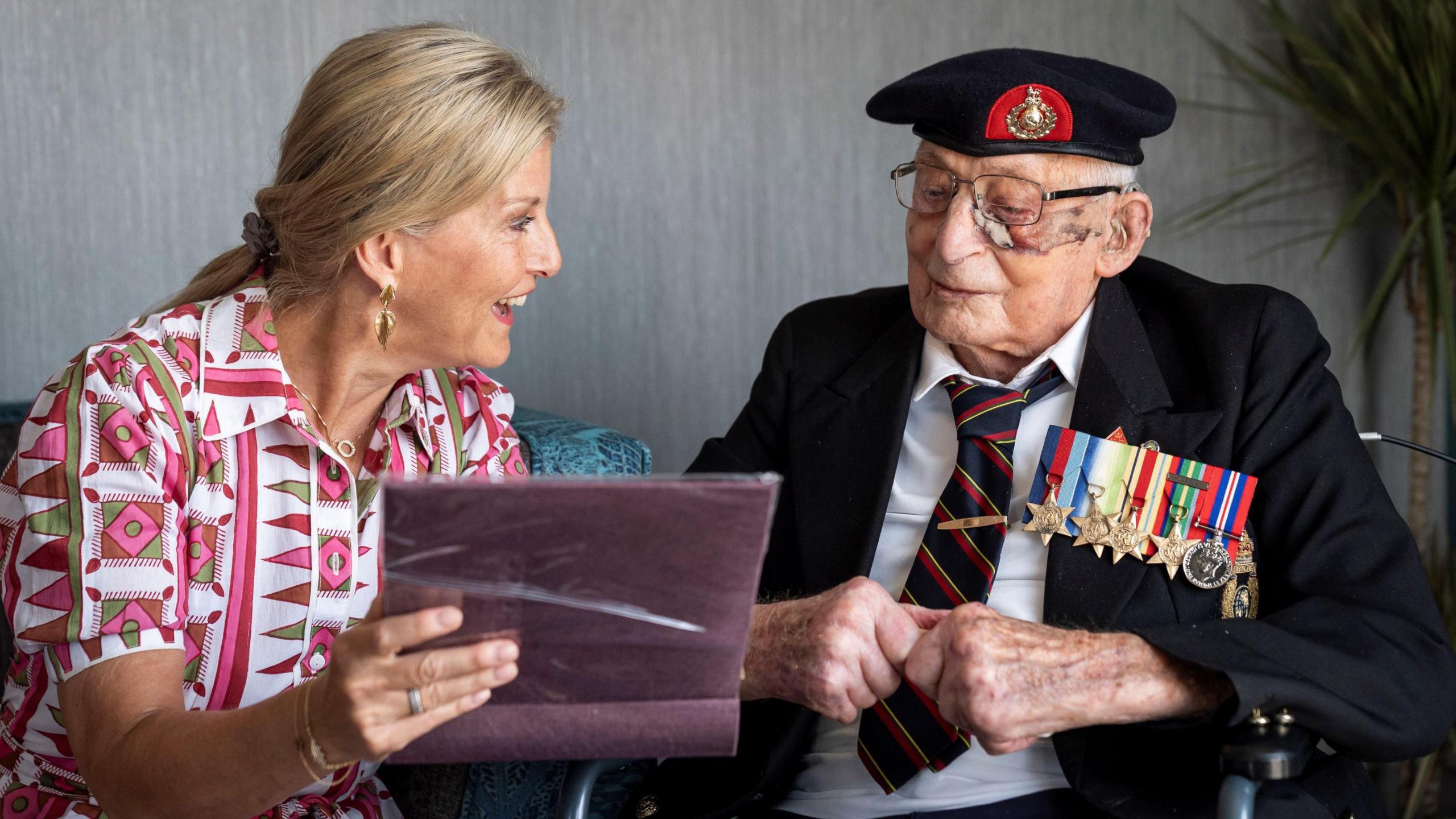 The Duchess of Edinburgh sits next to 105-year-old Royal Marines veteran James 'Jim' Wren. She is holding a photo album and going through it with him, She wears a dress patterned with pink and he is in uniform with multiple medals.