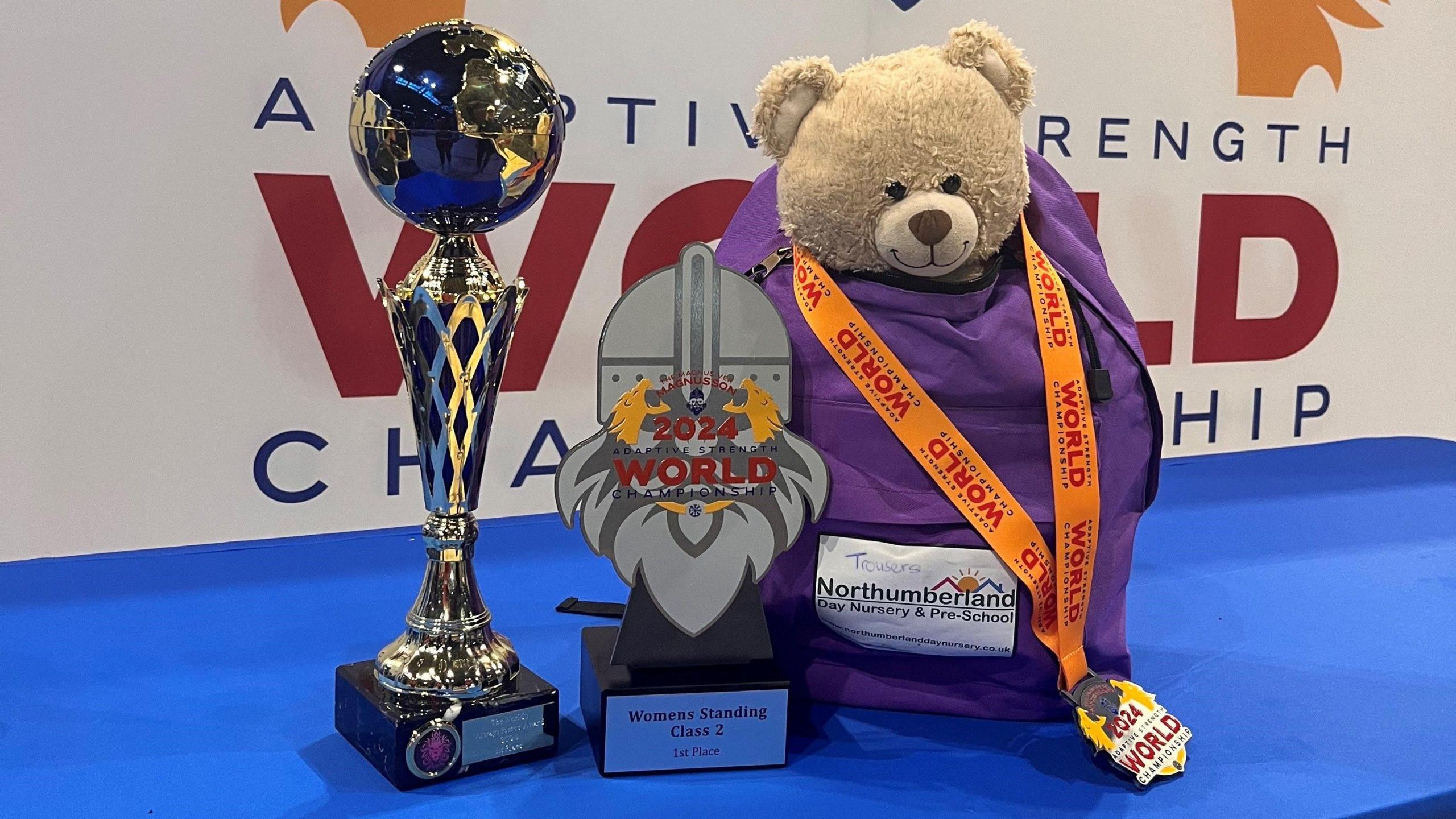 Two silver trophies are next to a purple backpack with a teddy bear poking out of it. There is a white background with red and blue writing on it