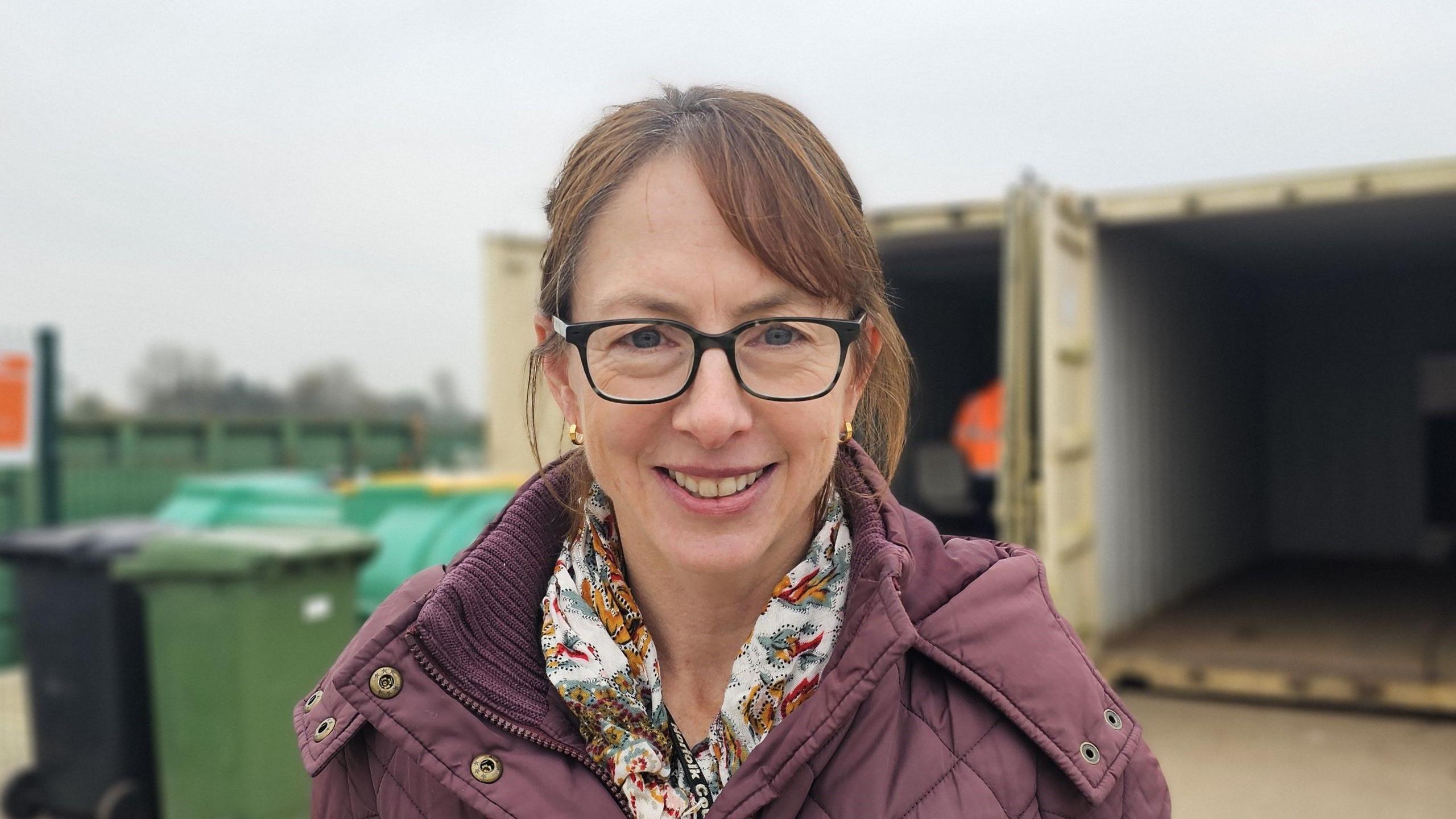 Kate Murrell standing in front of two large containers at a recycling centre. She has glasses and is wearing a light purple coat.