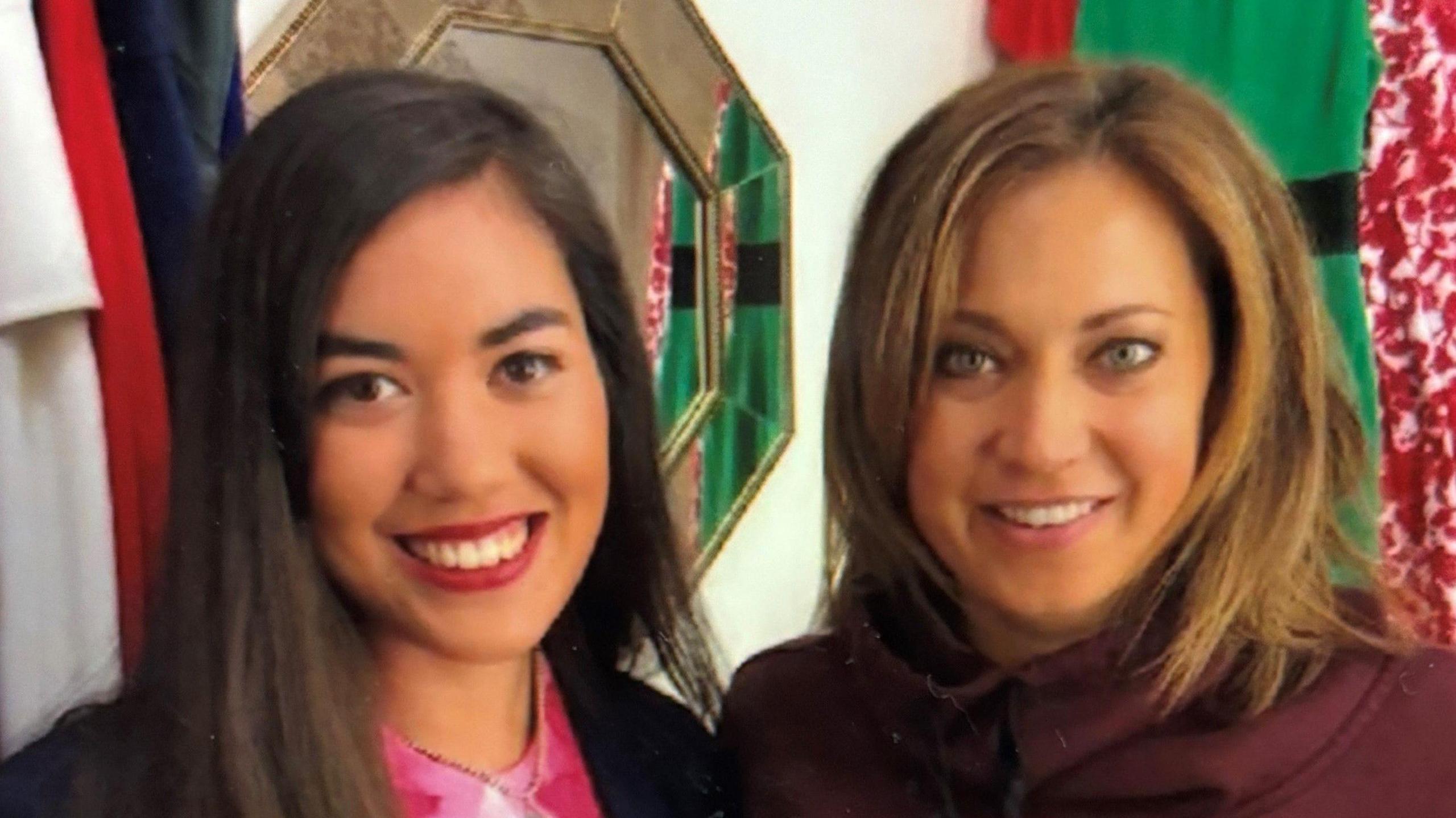 Two smiling women stood side by side. Girl to the left has dark brown hair and eyes. Woman to the right has caramel streaks with green eyes. Background busy with clothes hanging up.