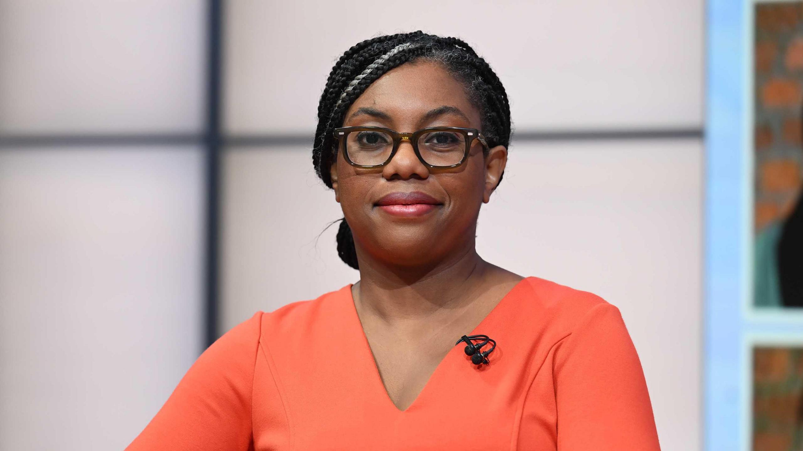 Kemi Badenoch looking straight at the camera. She is wearing a coral dress and glasses.