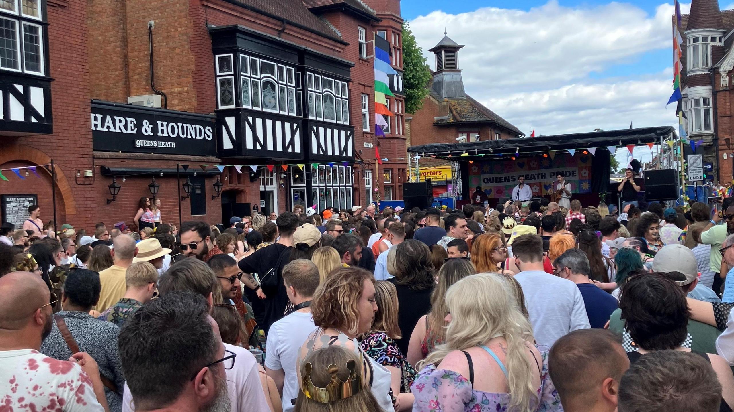 Picture shows hundreds of people on a street watching a stage at the far end where three musicans play. On the right hand side is the Hare and Hounds pub and its famous red brick and black and white facade. There is colourful bunting across the street and rainbow flags flying from buildings