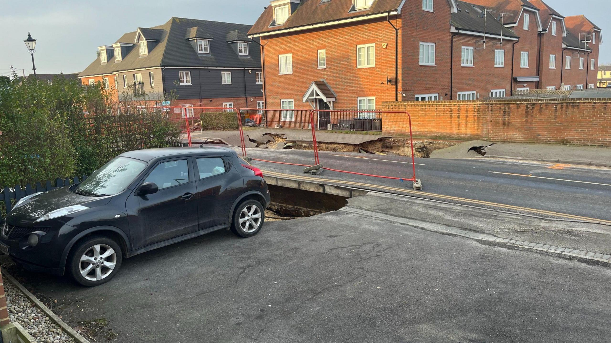 A dark car parked in a driveway of a property in Godstone, with the edge of a sinkhole reaching its rear wheels. The original, larger sinkhole is visible on the opposite side of the road, where both one lane of the road and part of the pavement have collapsed. There are a number of houses in the background.