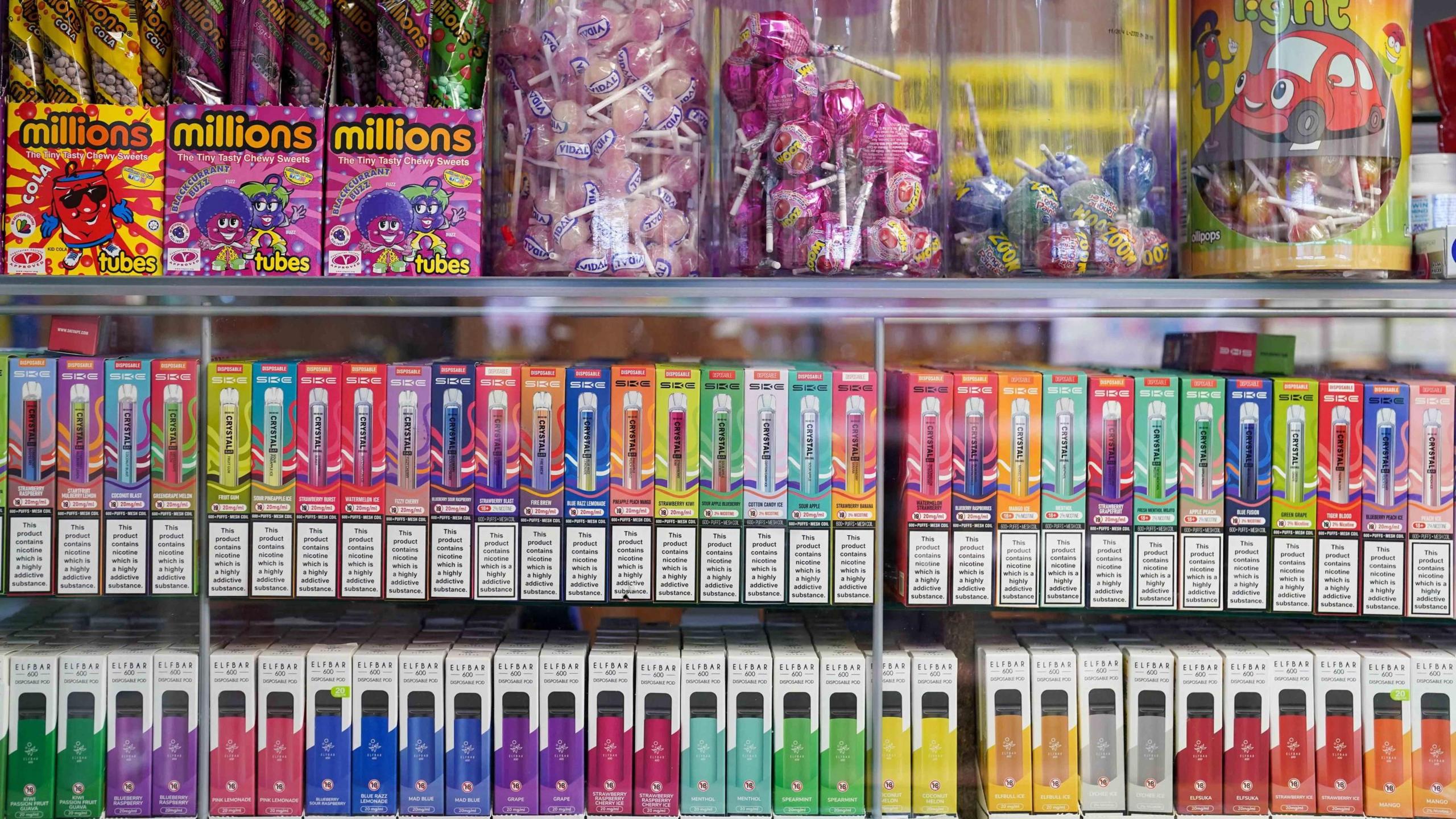 Disposable vapes are on display in a glass cabinet across two shelves. They are all different colours: pink, green, purple, yellow, blue, orange, red and white. On top of the glass cabinet are sweets. On the left are tubes of millions in orange and pink packaging and on the right are four boxes of lollipops. Some are bubble-gum pink whilst others are blue, green and red.