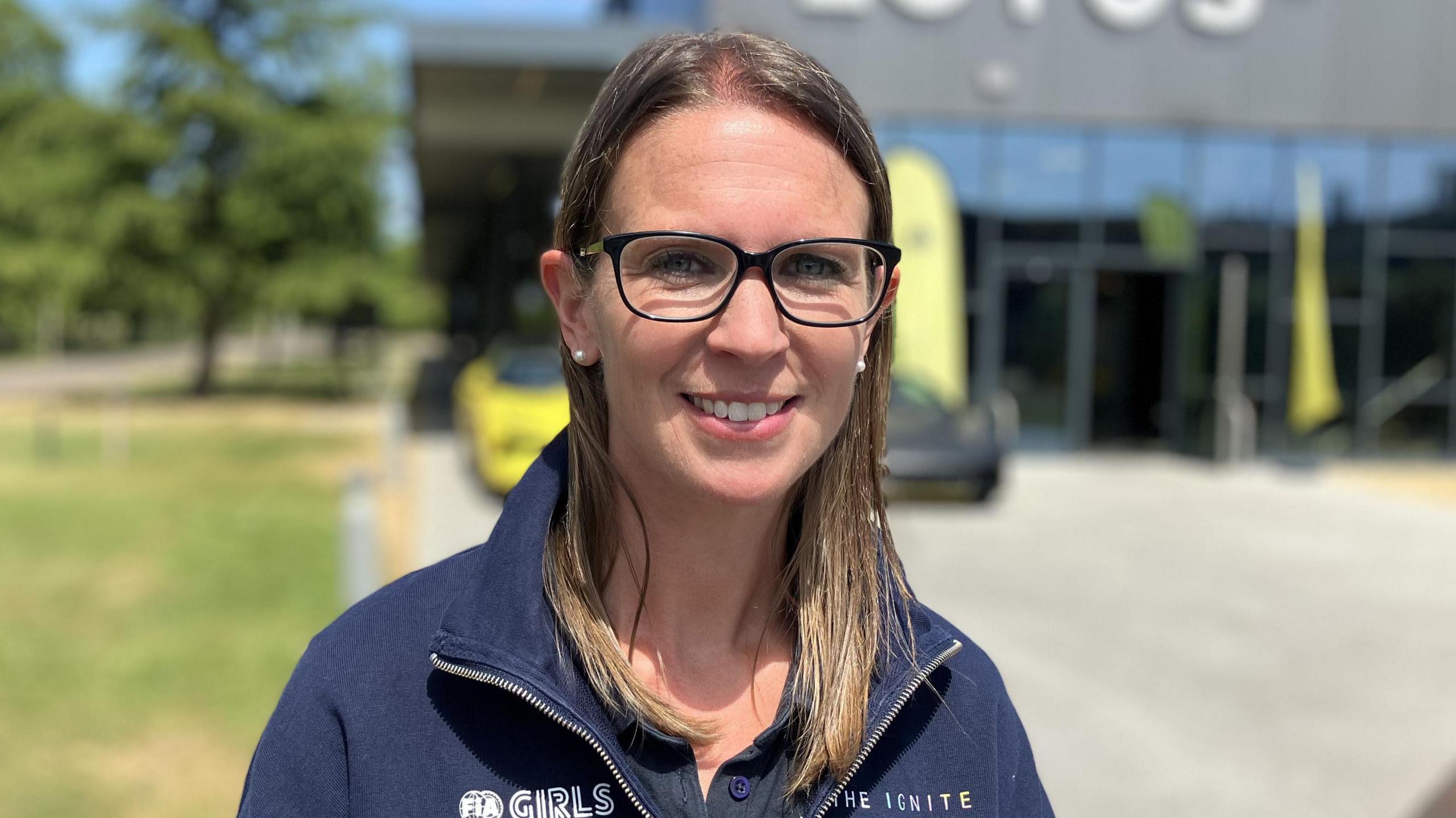 Jenny Fletcher is a woman with brunette straight hair who is standing outside the Lotus building and is looking directly at the camera and smiling.