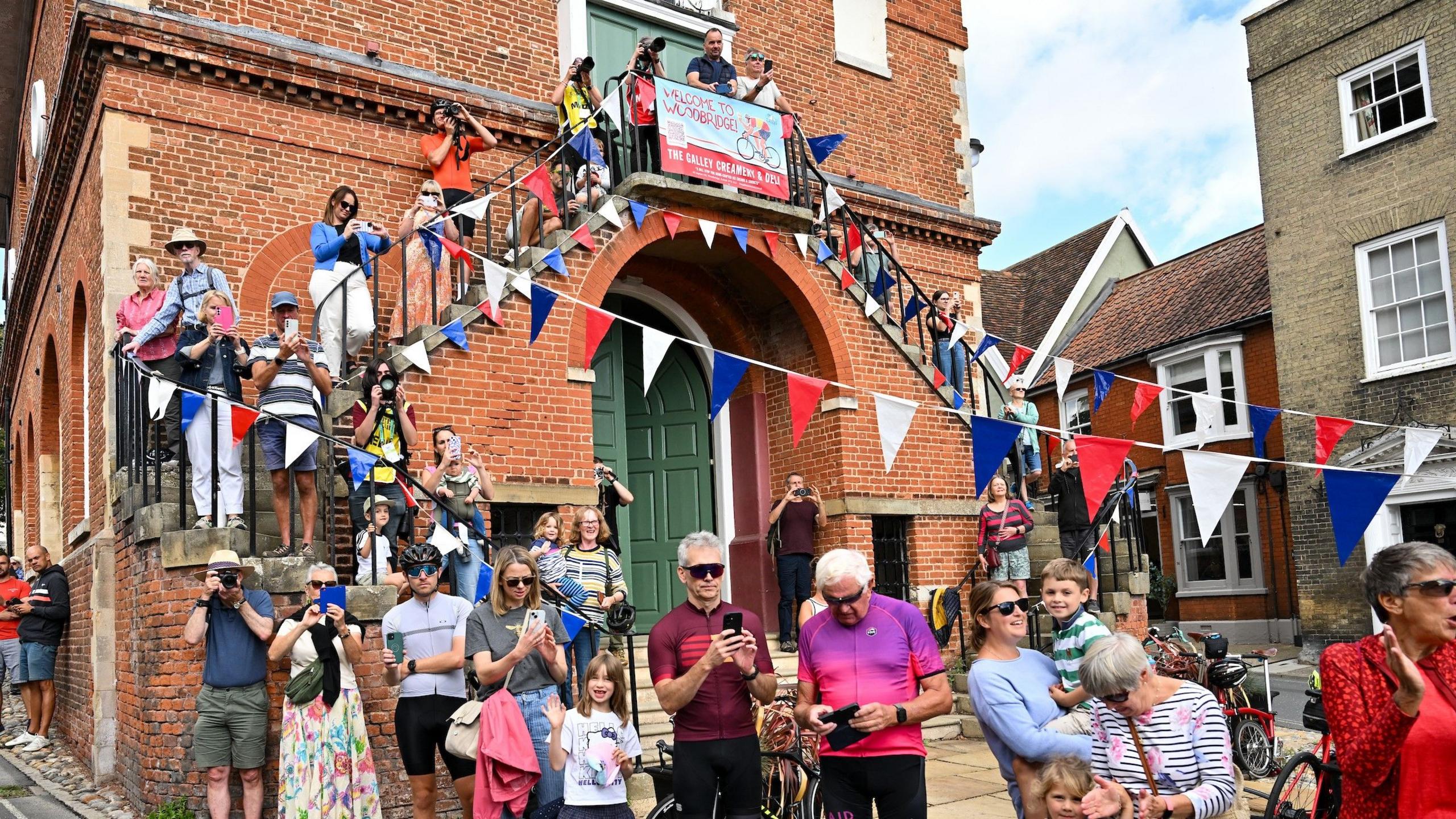 Crowds of people stand next to a road ready to cheer on the cyclists. Red, white and blue bunting has been put up along part of a building and above the road.