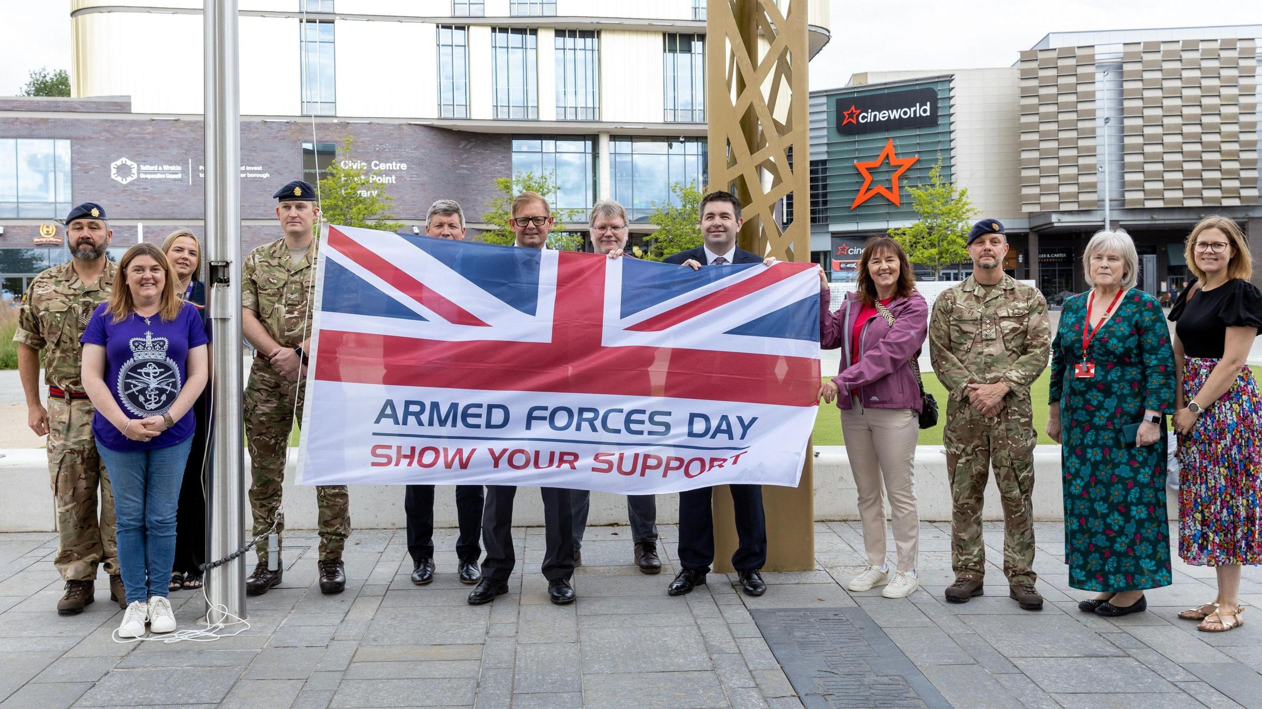 A group of people, including three men in camouflage style military uniform are standing in a line. In the middle, they are holding a large flag that has half a union jack on it, with the words "armed forces day, show your support" on the bottom half.