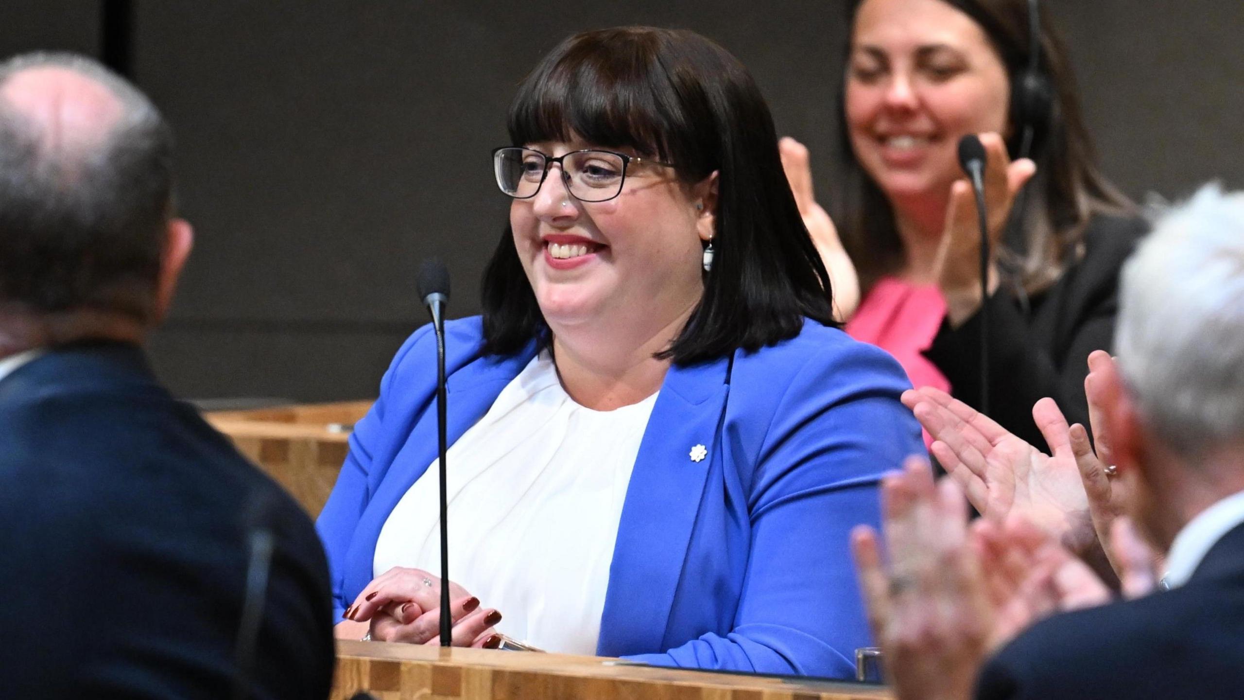 Kerry Ferguson - menyw gwallt du canolig mewn blows wen, siaced las a sbectol liw tywyll - yn gwenu yn siambr y Senedd ym mis Mai ar ôl cael ei hethol yn Ddirprwy Lywydd