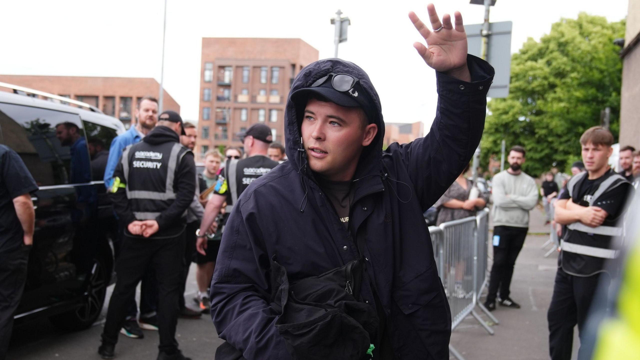 Liam Ó hAnnaidh, who is wearing a dark raincoat with the hood up over a black cap and sunglasses, waves to crowds outside Glasgow's O2 Academy. Behind him are his security team standing in front of a dark vehicle.