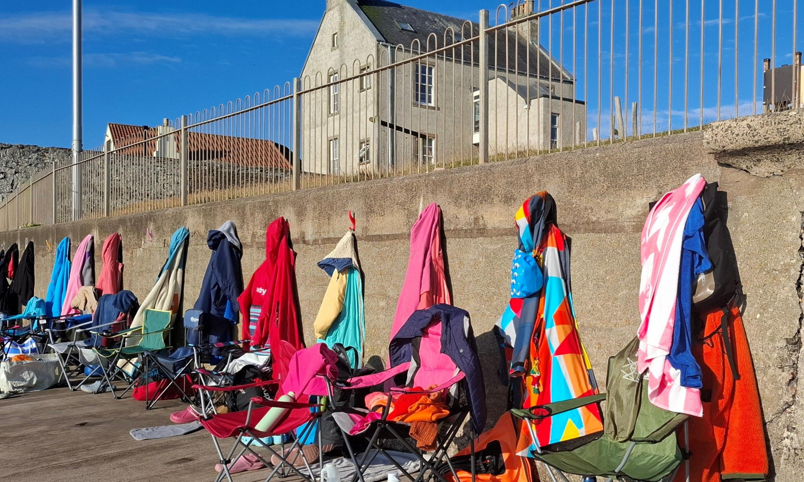 A row of brightly-coloured towels and robes hang along the beach wall like a rainbow at Cellardyke Tidal Pool, camp chairs on the beach also hold swimmers' things.