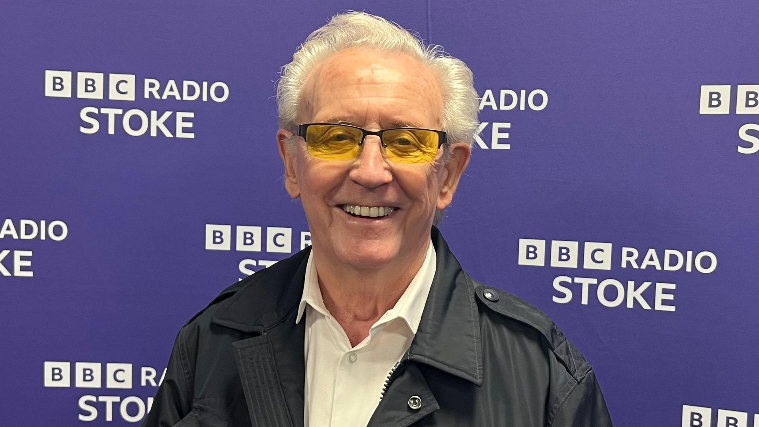 Tony Christie, with short white hair, yellow tinted glasses, a black jacket and a white shirt, smiles while he stands in front of a purple background that says BBC Radio Stoke in white letters.