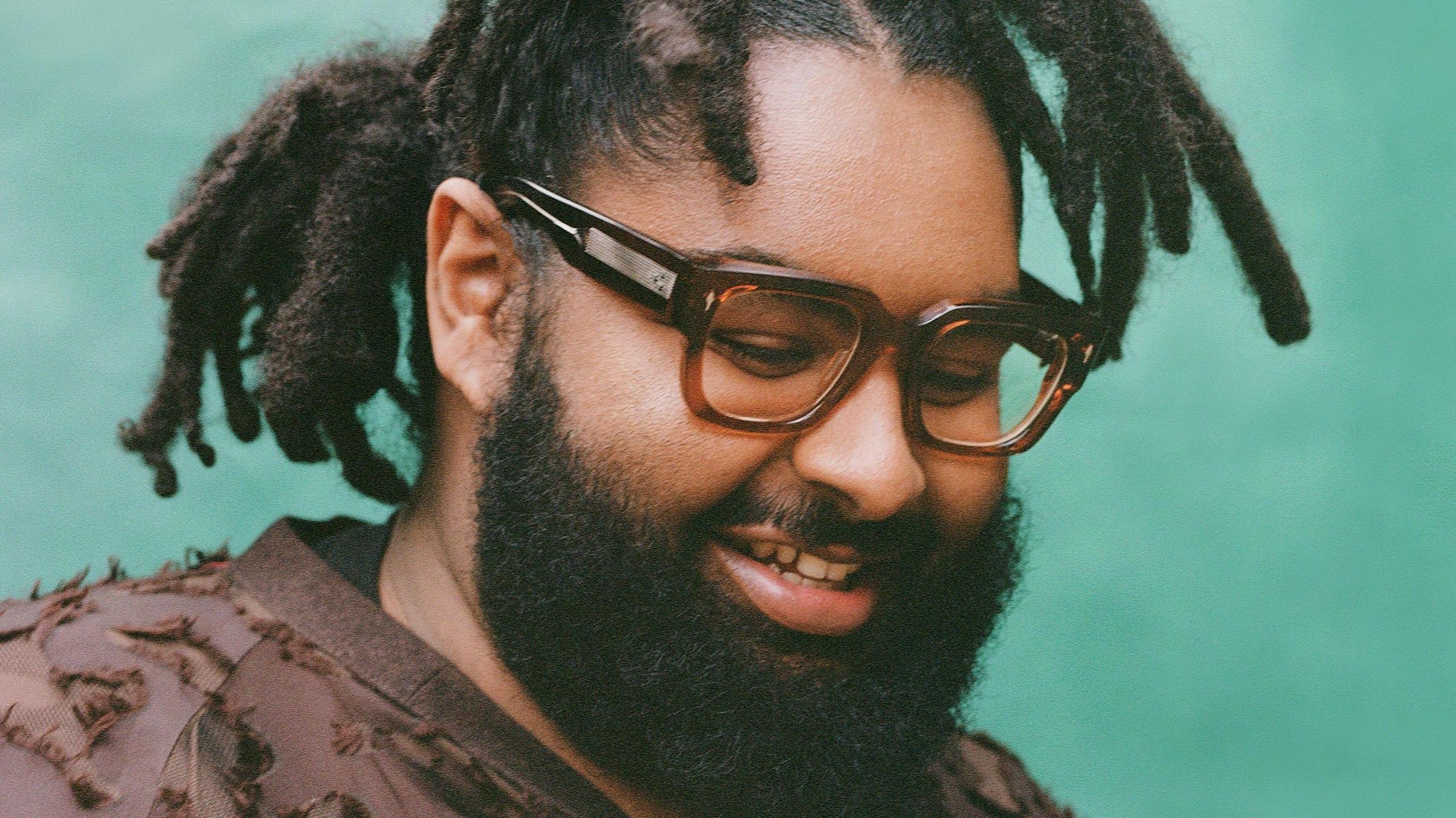 Elmiene pictured looking down and smiling. He wears a brown top and square tortoiseshell-rimmed glassed. He has a thick beard and wears his hair tied back and stands in front of a blue backdrop. 