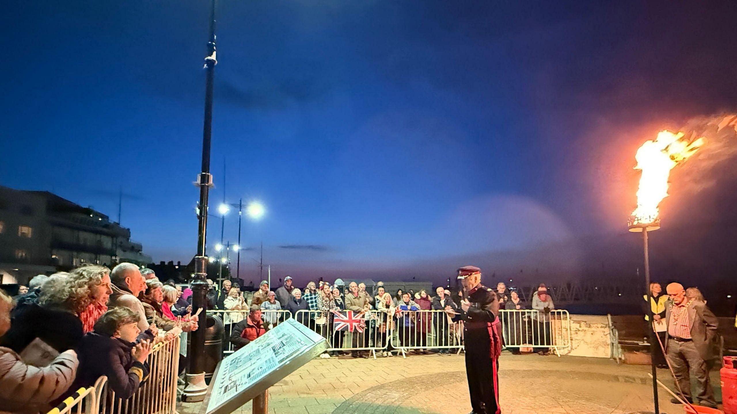 Hundreds of people gathered behind barriers at the beacon ceremony. To the right of the photo is the burning beacon - a metal basket of burning wood on top of a pillar.
