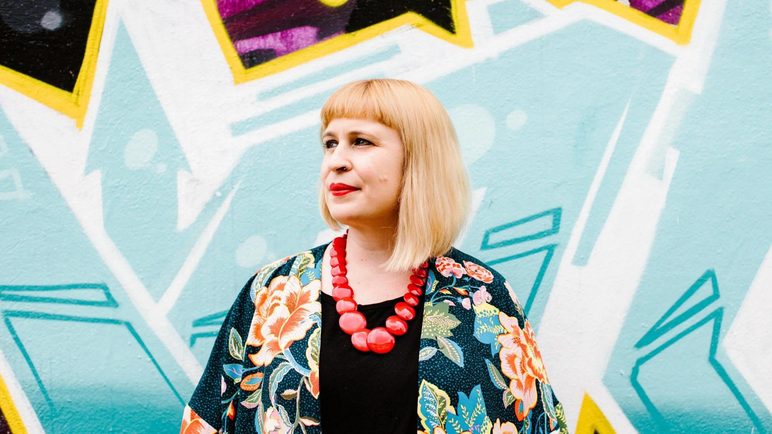 A woman with blonde hair and a floral top is standing in the centre of frame in front if a blue wall with a funky pattern.