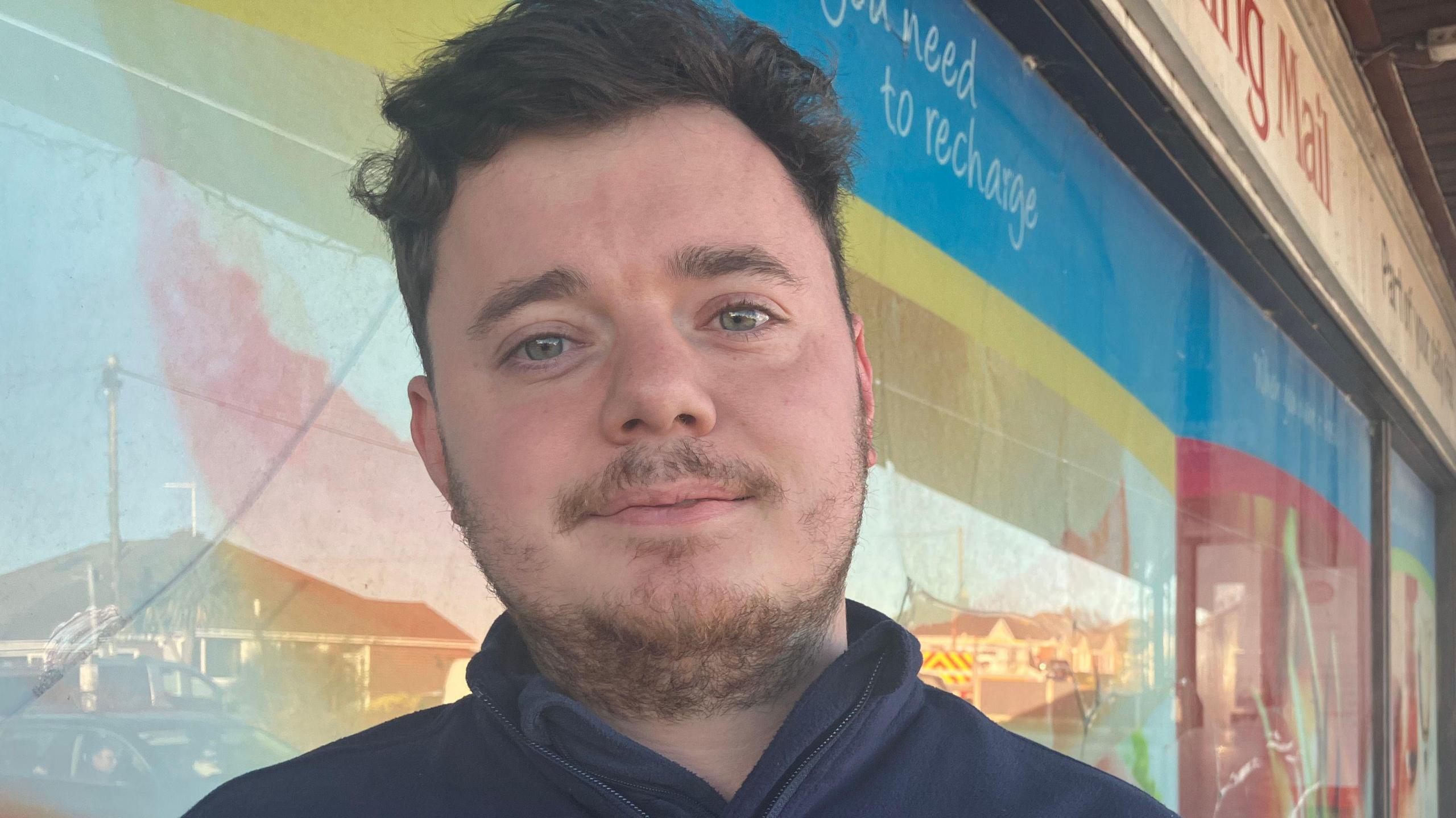 Daniel Copper, with short brown hair and a short brown moustache and beard, wearing a navy fleece, stands outside his convenience shop. The window is covered in a brightly coloured design and part of a large sign advertising a newspaper is visible behind him.
