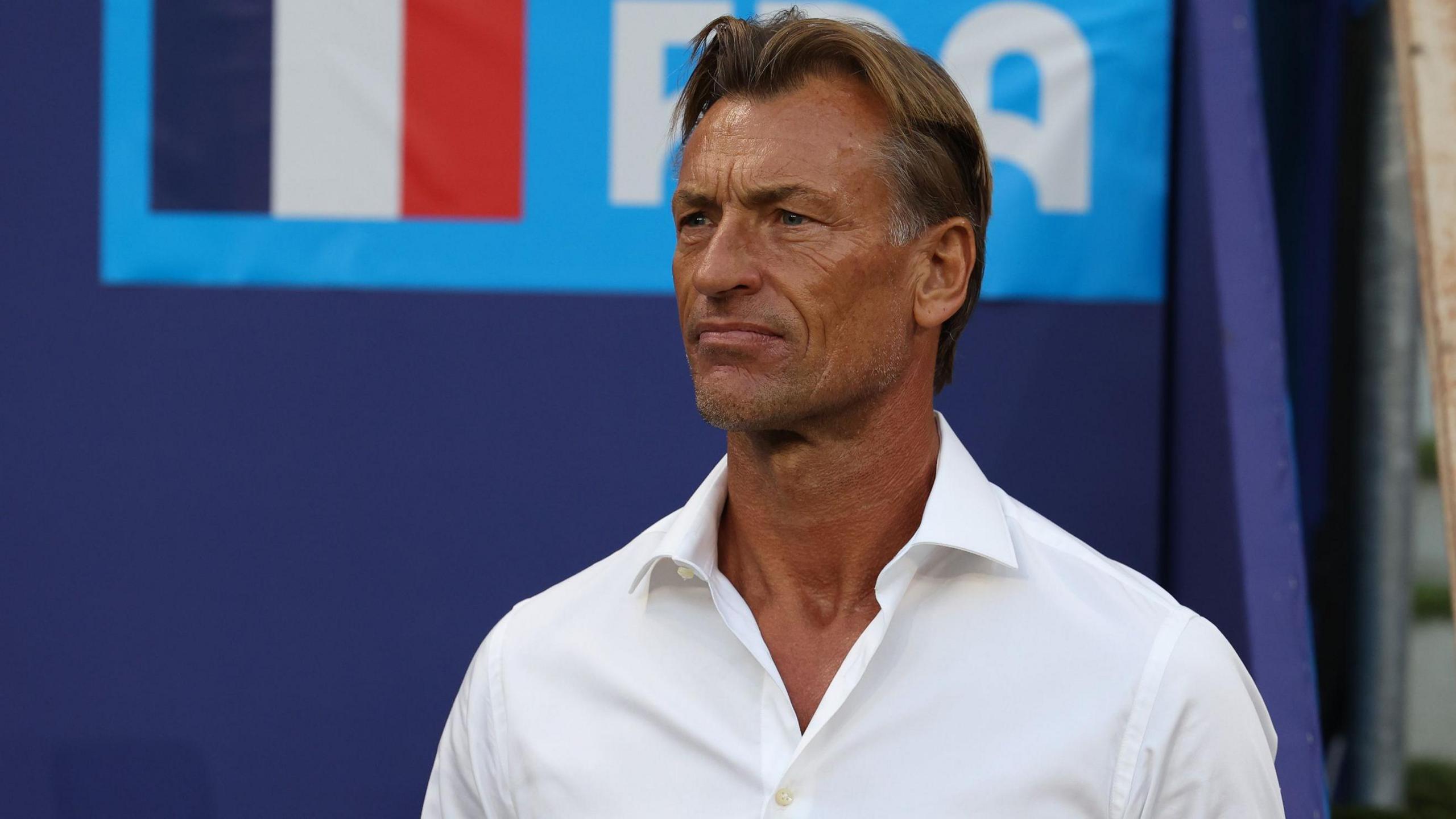 Herve Renard, visible from chest up, wears a white shirt unbuttoned at the neck as he stands impassively on the touchline at a football match. A purple hoarding bearing the French flag is out of focus in the background