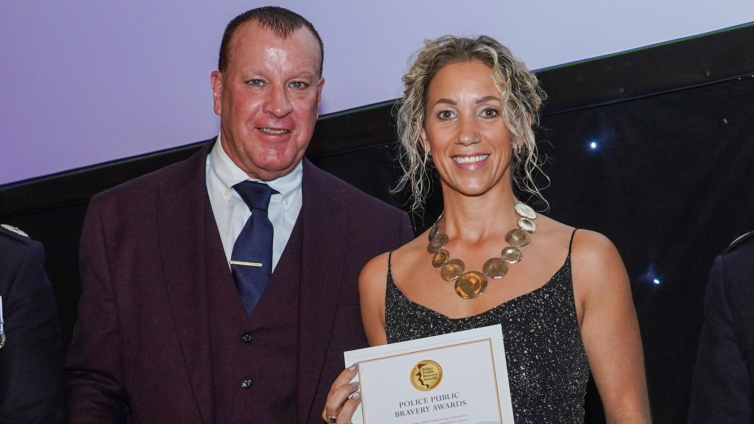A man in a suit standing next to a woman in a dress who is holding a police bravery certificate. She is wearing a necklace which is made up of a series of round bits of jewellery. They are both smiling into the camera.
