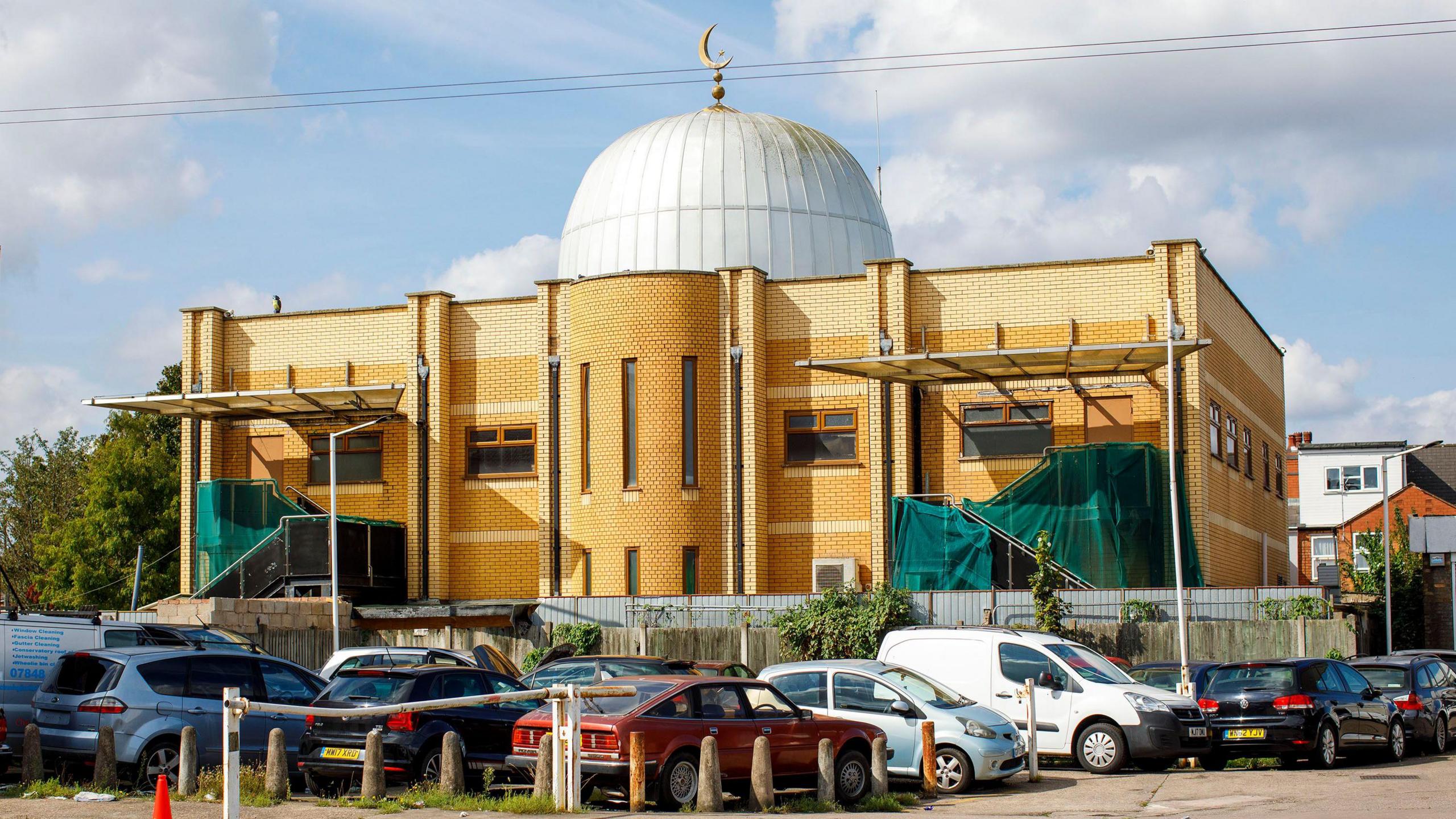 A large gold coloured mosque with a white dome on the top of it. There are cars parked in front of it.