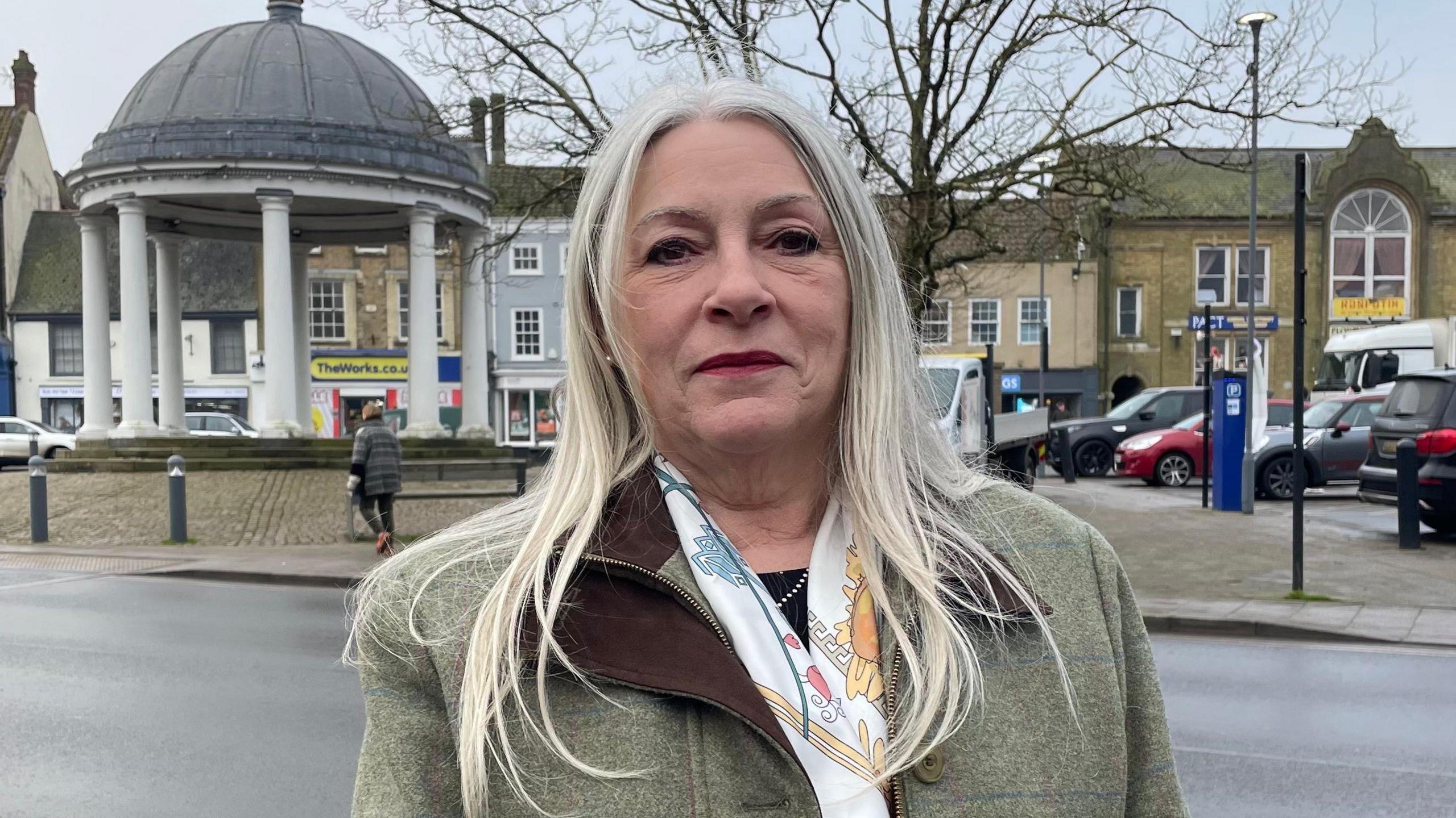 Kay Mason-Billig looking direct to camera and standing in Swaffham's market square. She has straight, grey hair that reaches below her shoulders and is wearing a green, tweed overcoat and a silk scarf, that is mainly white, with patches of blue and yellow. Behind her and in the left of the shot is a rotunda with a grey, domed roof and white columns, with three steps leading up to it. Over her other shoulder is a car park, where several vehicles are parked. Further in the distance are buildings and shop fronts.