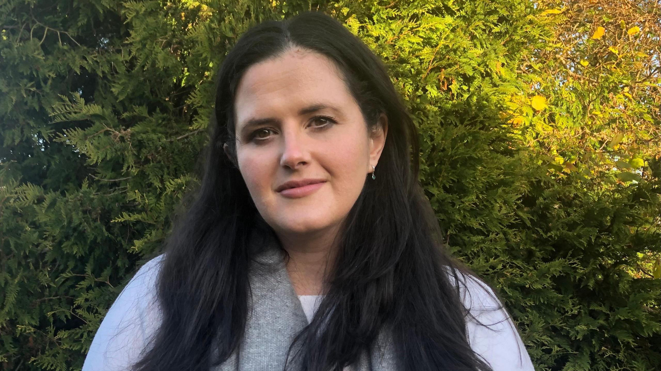 Claire Sudgen MLA standing in front of an evergreen hedge like tree. She has long dark hair and is wearing a white to and grey scarf. 