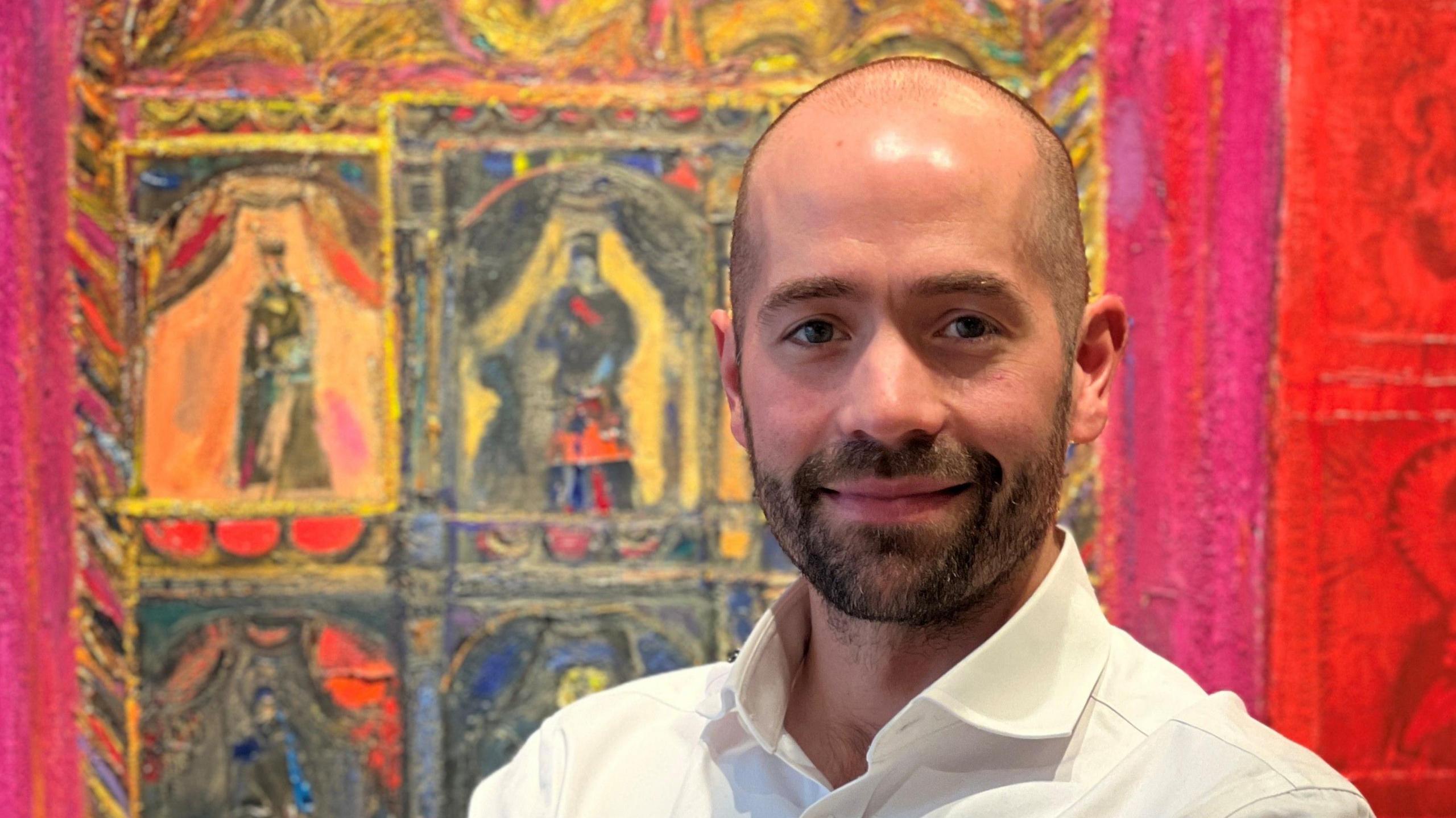Portrait of a man in a white shirt, smiling slightly while standing in front of a vivid, colourful painting with theatrical figures and bold red and gold tones.