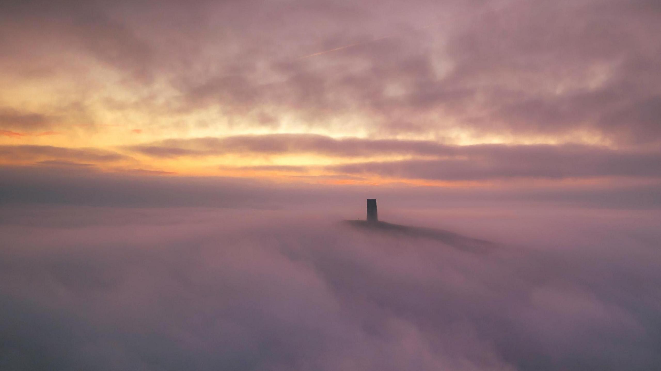 Glastonbury Tor can be seen in a sea of fog. It is just barely poking out of thick, white fog as the sun is coming up behind the hill.