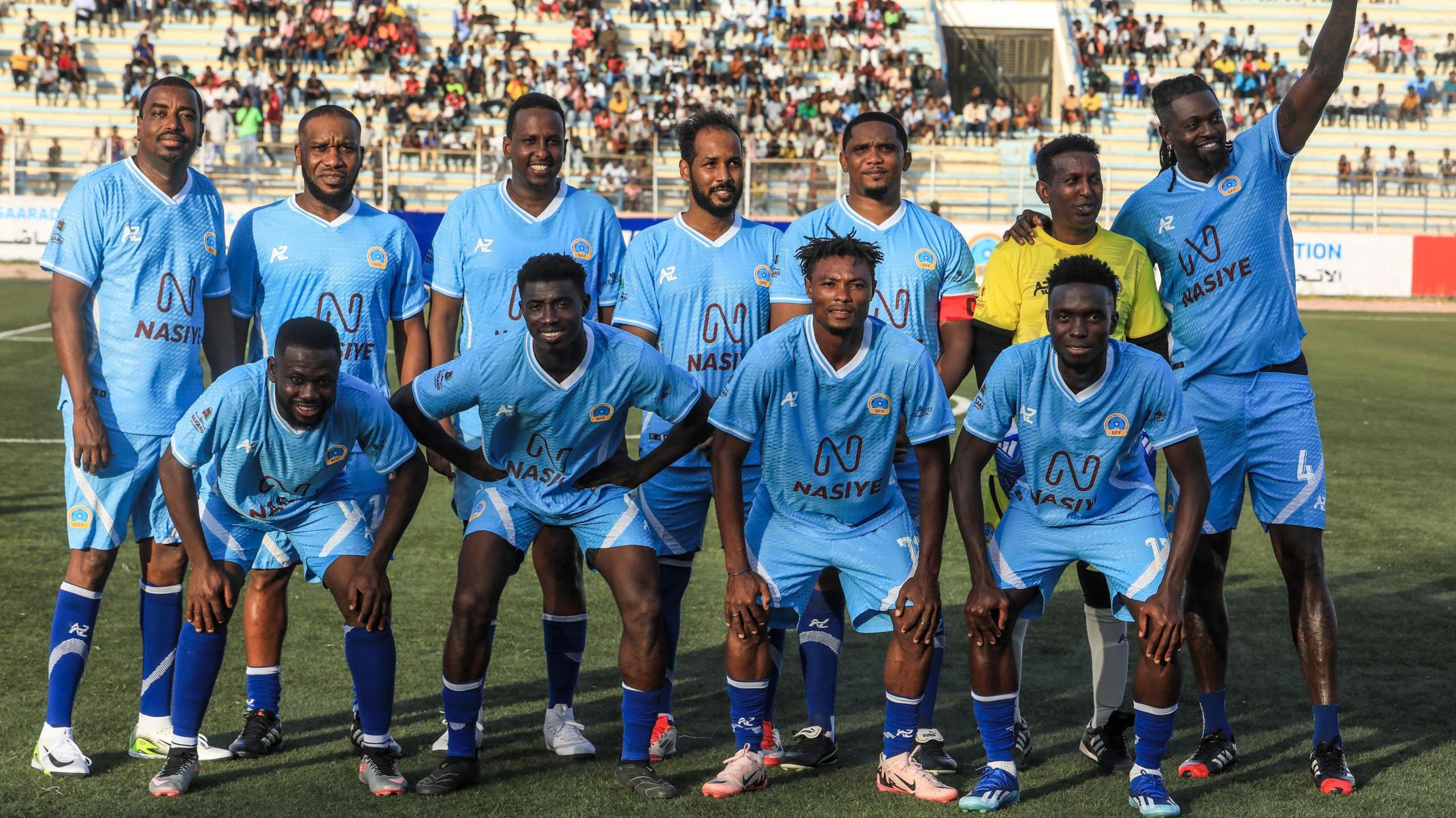 A football team wearing light blue kit is pictured before a game, with Emmanuel Adebayor on the right-hand side of the back row waving by raising his left arm while having his other arm around a goalkeeper in a yellow top