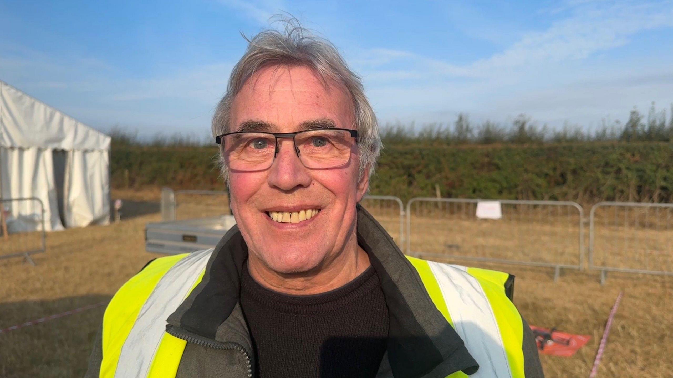 A man, who has grey hair, is wearing black glasses, a black top and a yellow high-visibility jacket with silver reflective strips. He is standing in a field with a white tent, metal railings and greenery behind him.