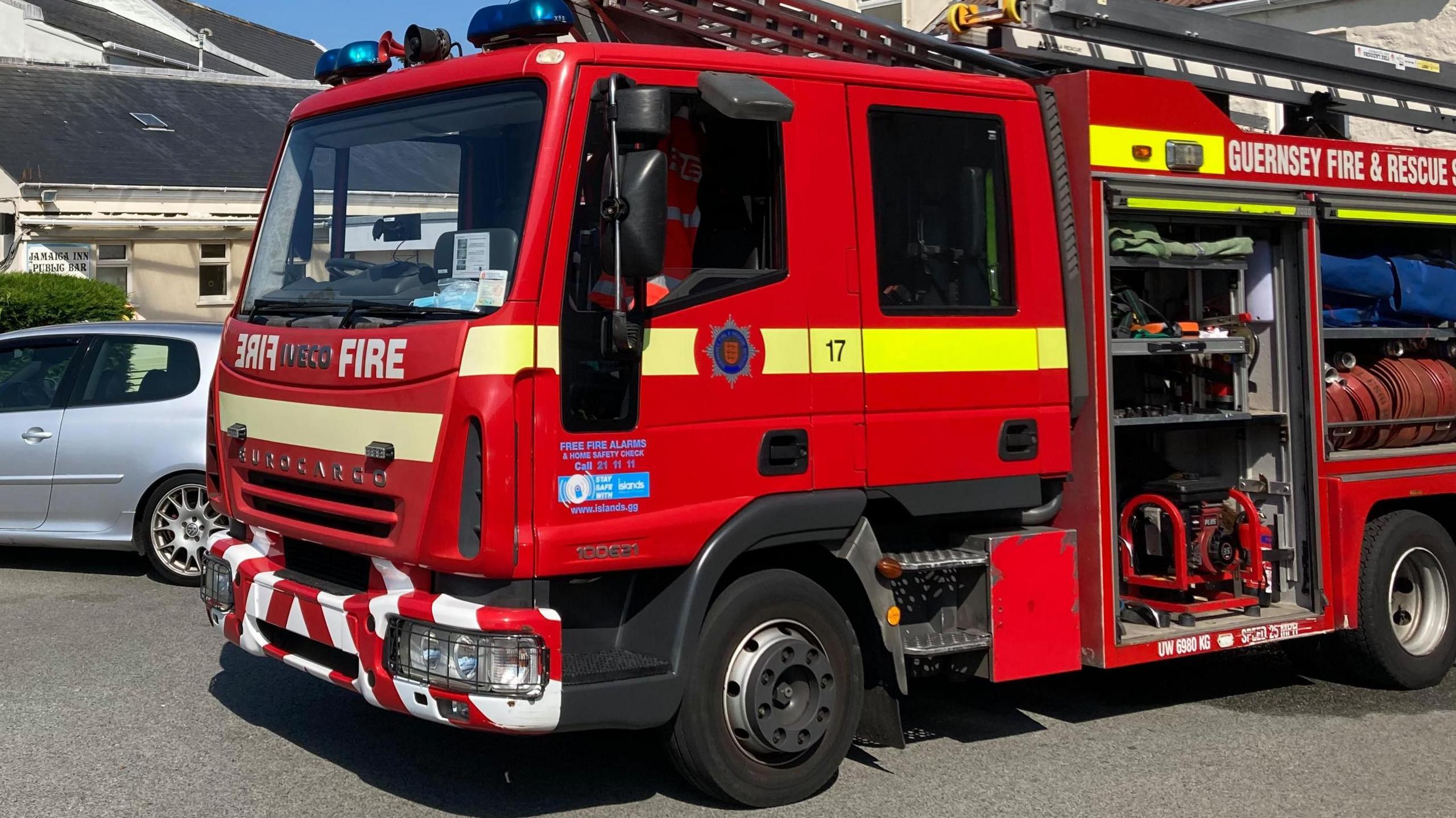 A red Guernsey fire and rescue fire engine outside in a built up area with blue skies. The side panels where the hoses are kept is open. There is a silver car next to the truck. 