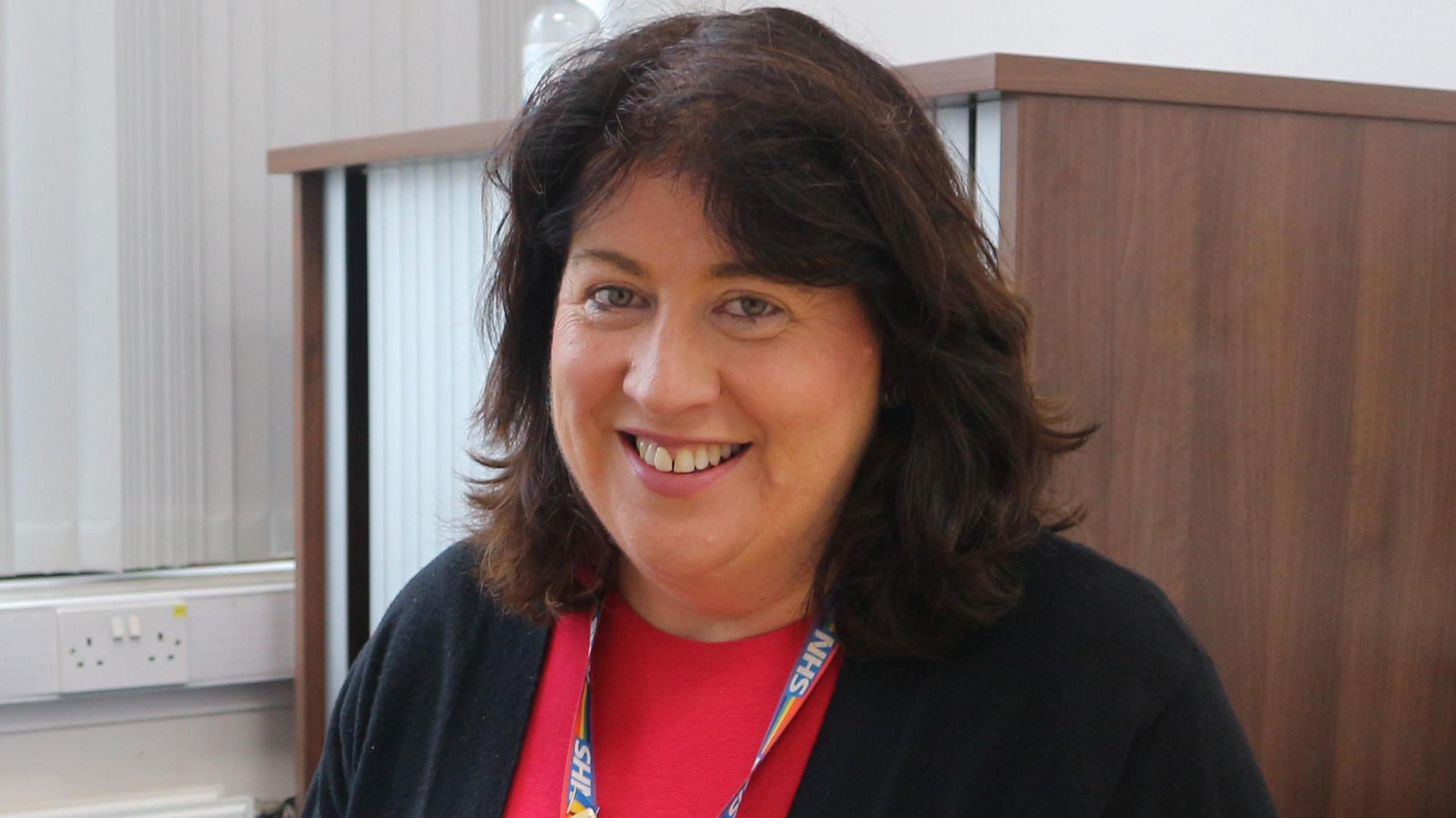 Siobhan Harrington smiles at the camera. She has shoulder-length, brown, wavy hair and wears a black cardigan over a red top with an NHS lanyard.