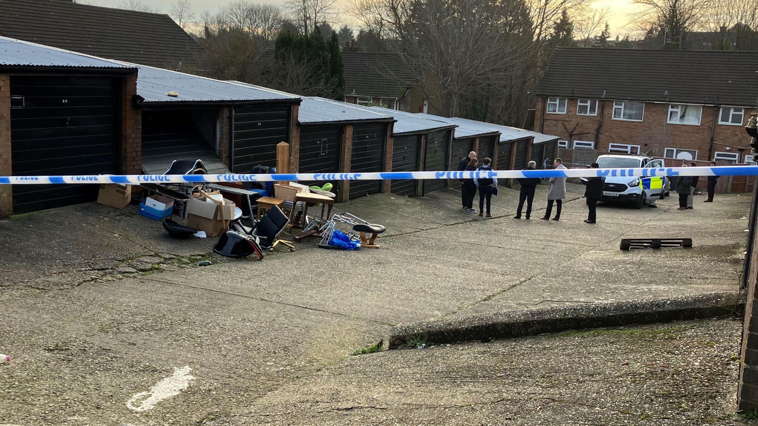 Police tape prohibits entry to some garages. Household items are stacked up outside one of the garages, and a police van and several uniformed and plain-clothes staff are in the distance. There are houses beyond the van