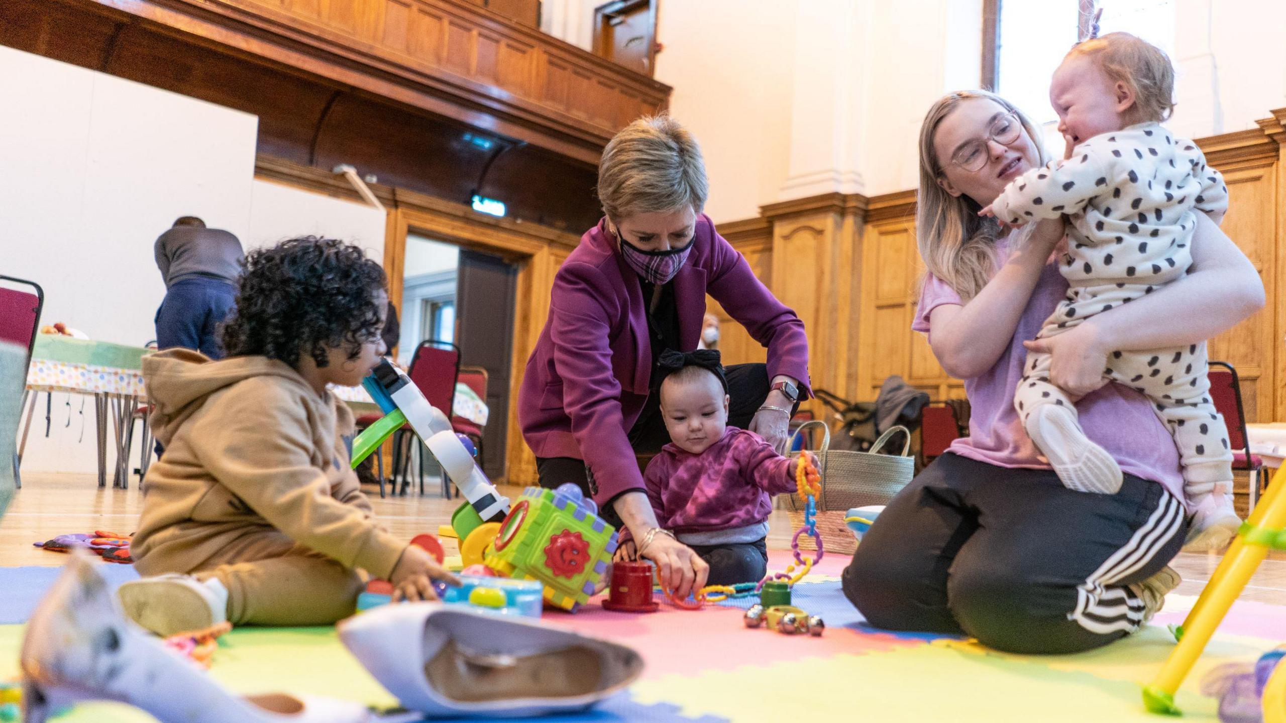 Nicola Sturgeon playing with children at a day care event - she has a face mask on and is is wearing a purple jacket. She is sitting down. Two children are also sitting down and playing with toys on the ground. Another woman, with glasses, blonde hair and a pink top, is also sitting down and is talking to  another child, in her arms. 