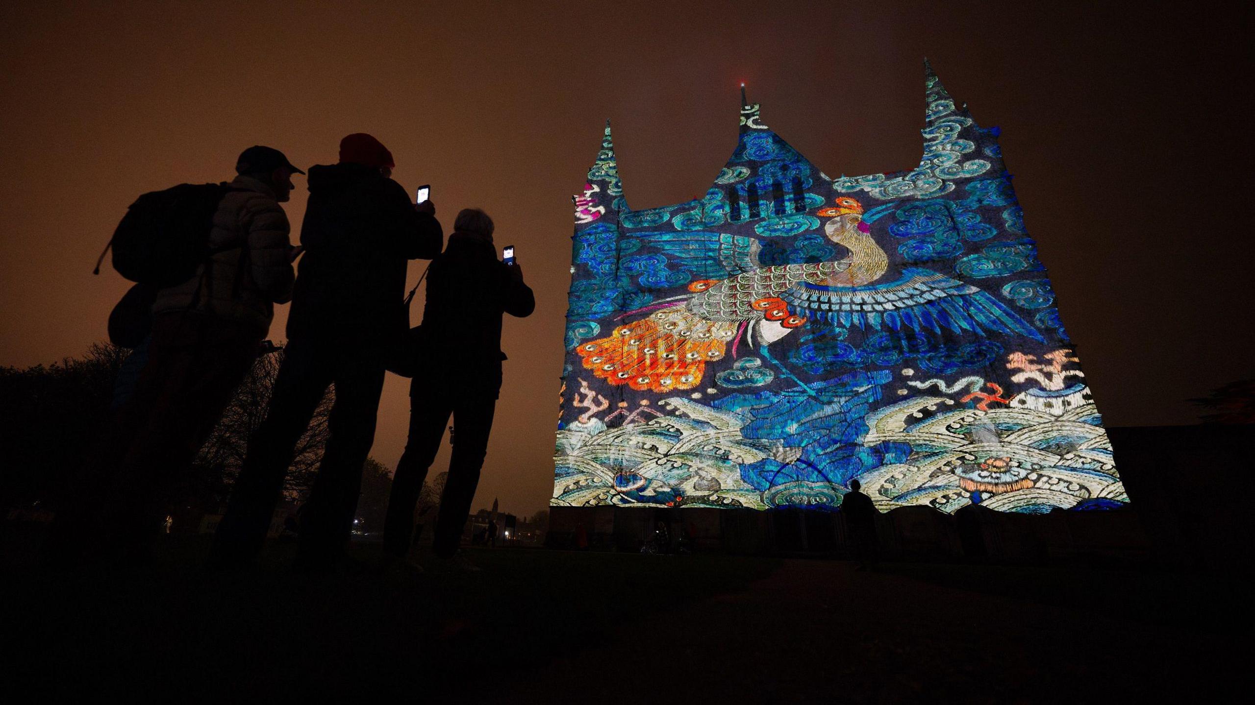 Three people taking photos on their phones of the light display projected on Salisbury Cathedral on a dark night. The light display features intricate patterns illuminated on a wall, with a colourful bird, possibly a peacock, featured in the middle.