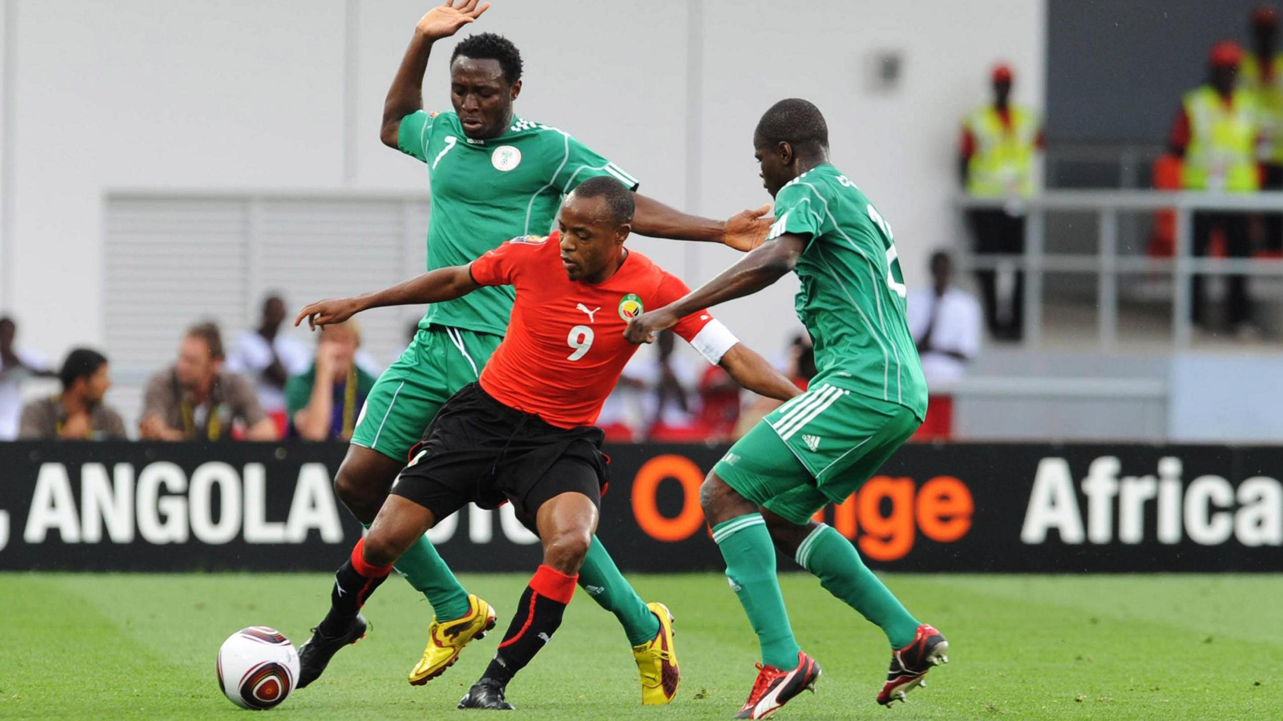 Manuel Bucuane, wearing a red Mozambique shirt, black shorts and black socks and red trim, attempts to control a ball while under pressure from two Nigeria players wearing an all-green kit. In the background several people are seen out of focus behind a black advertising hoarding