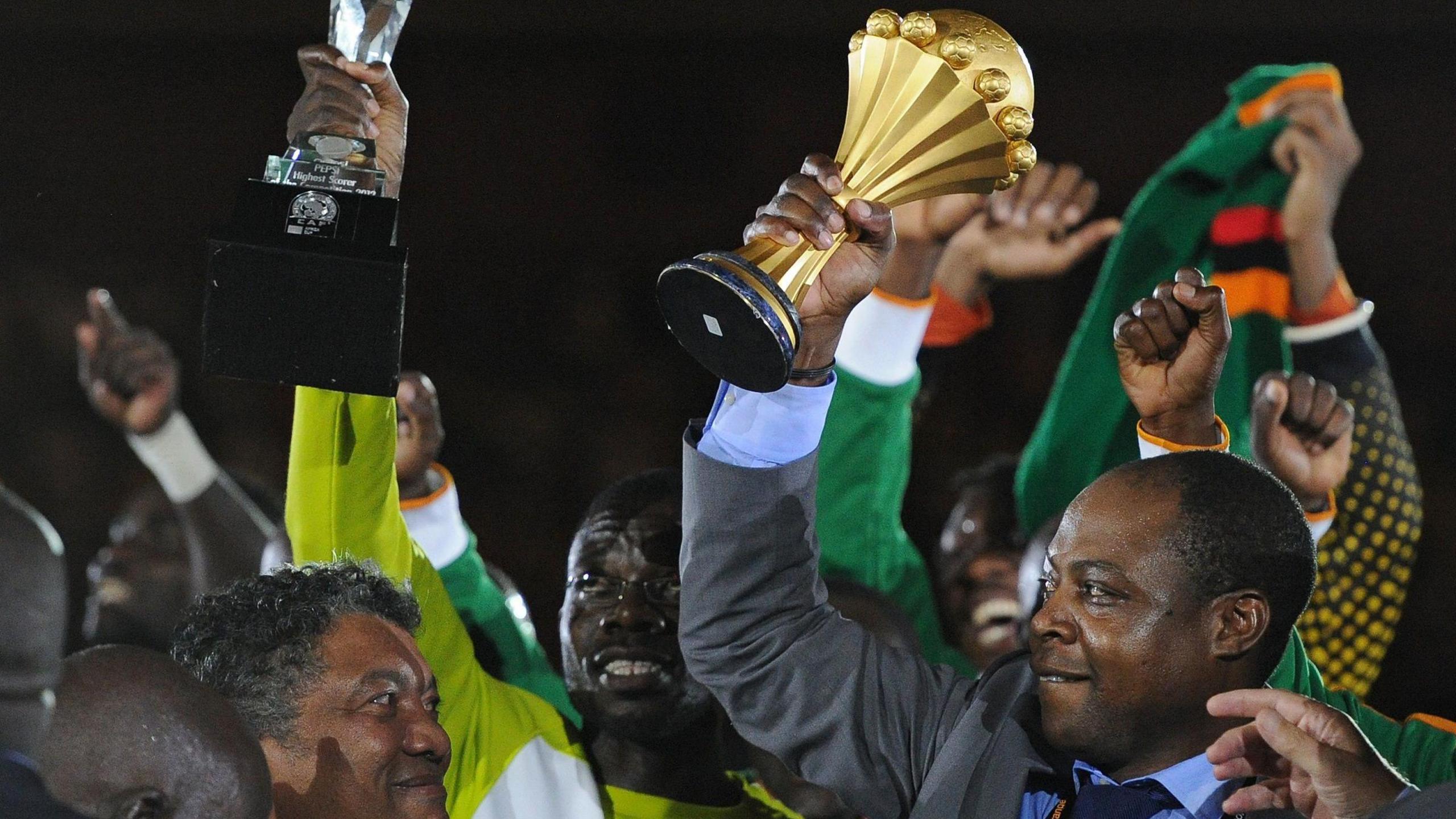 Kalusha Bwalya in grey suit holds up the Afcon trophy as others celebrate around him.