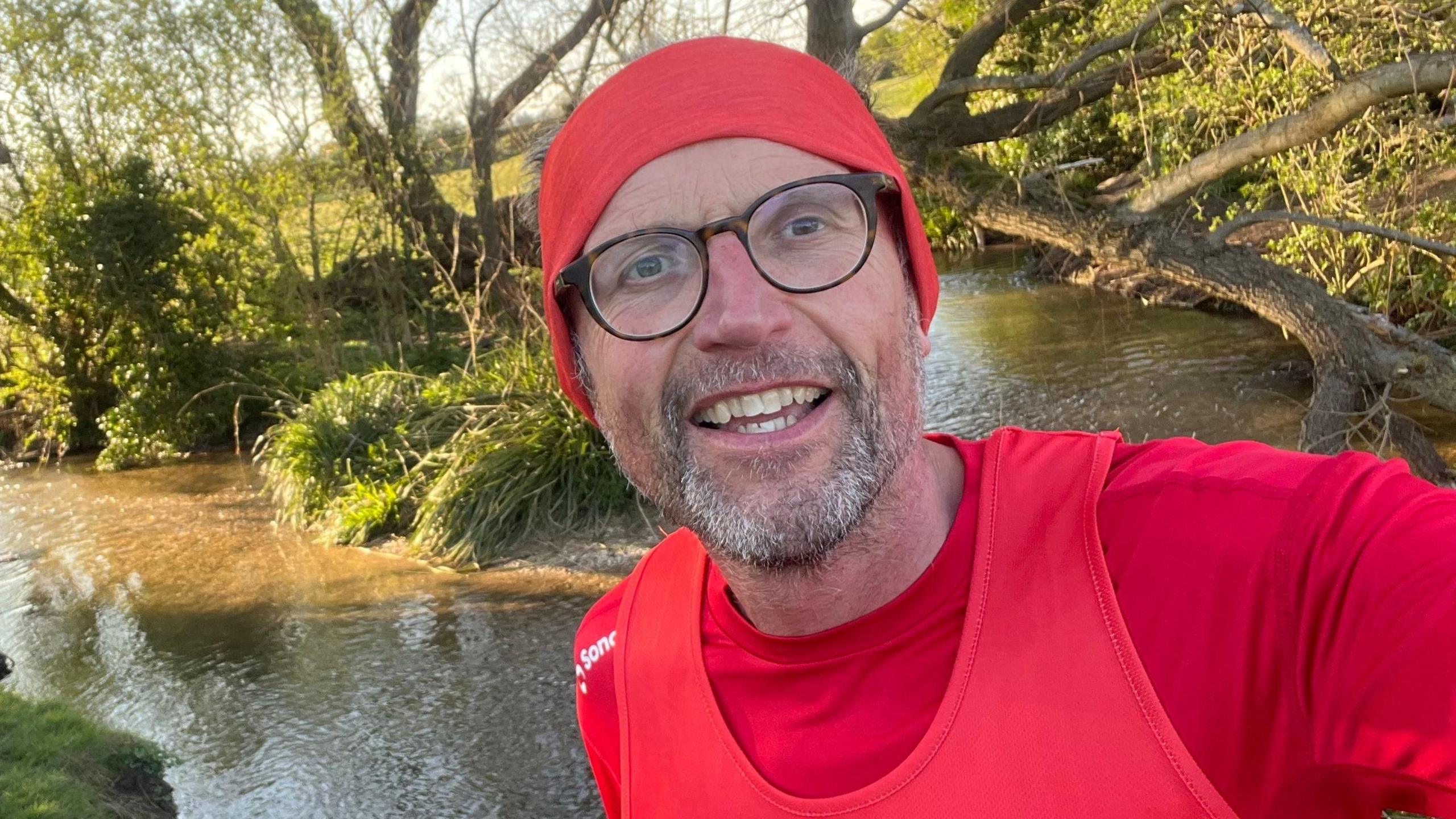 Rich Williman is at the centre of the photo. He's wearing a red running top and matching red headband. He's wearing glasses and is smiling. In the background there is a river and lots of plants and trees.