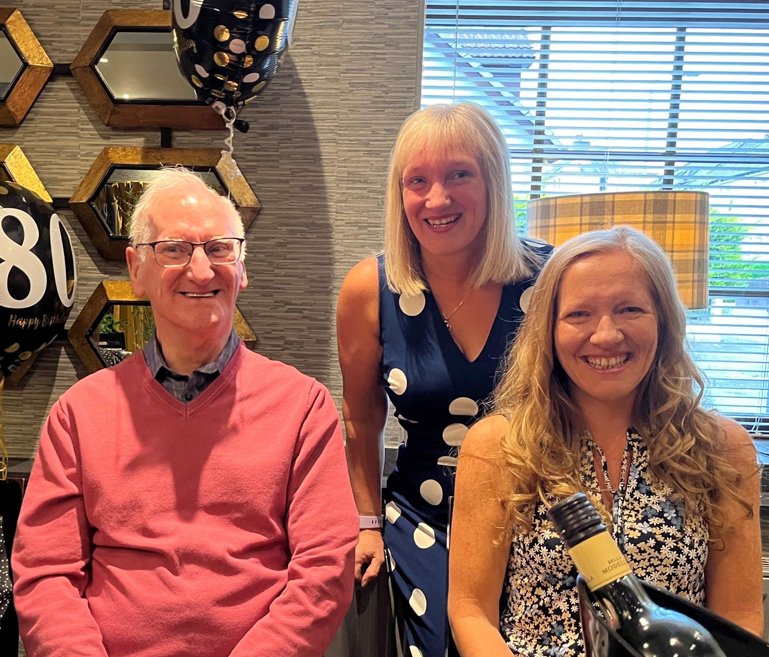 A man in a pink jumper with blue shirt underneath with grey hair and glasses sits next to two women - both with blonde hair - one is in a dark blue dress with white spots and the other in a floral dress. They are in a restaurant with a bottle of wine in the foreground and a balloon with the number 80 on it in the background.
