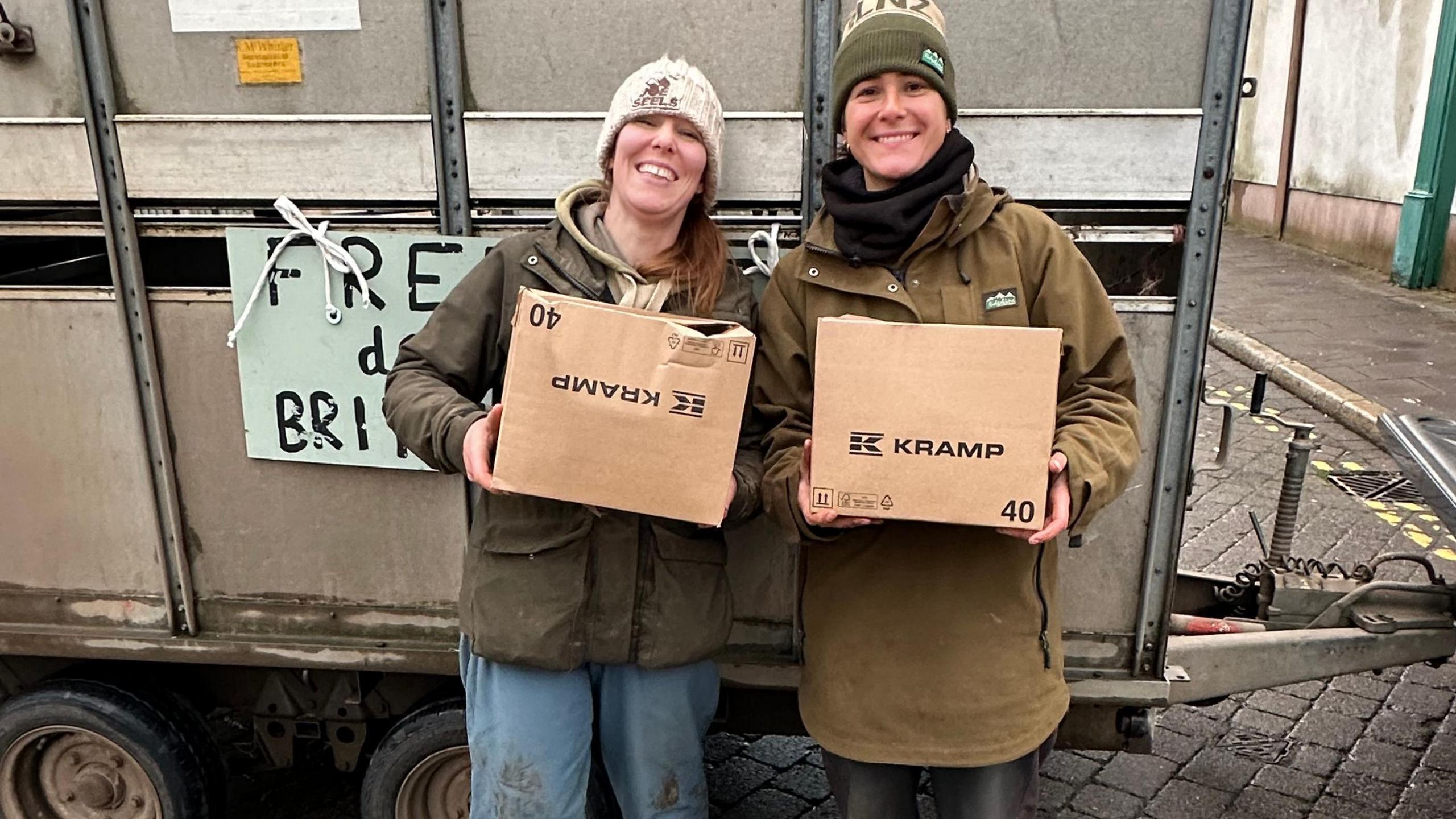 Two farmers, Charlotte Ashley and Rebecca Wilson, stand together holding two of the food donation boxes, which are cardboard. Both woman wear bobble hats, wax coats and jeans.